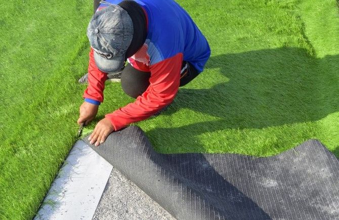 A person in a red and blue shirt cuts a piece of artificial turf with a utility knife to fit it over a grave site.