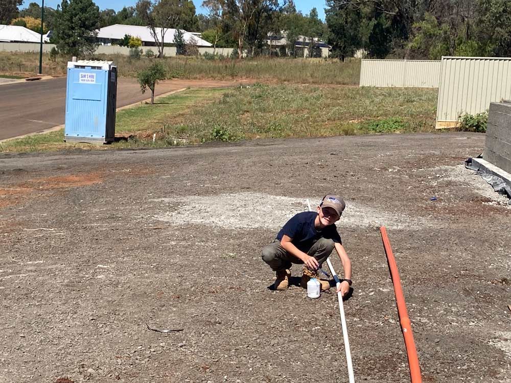 Person Kneeling on Gravel Ground, Painting a Line Near Orange and White Poles — Rohr Electrical in Dubbo, NSW