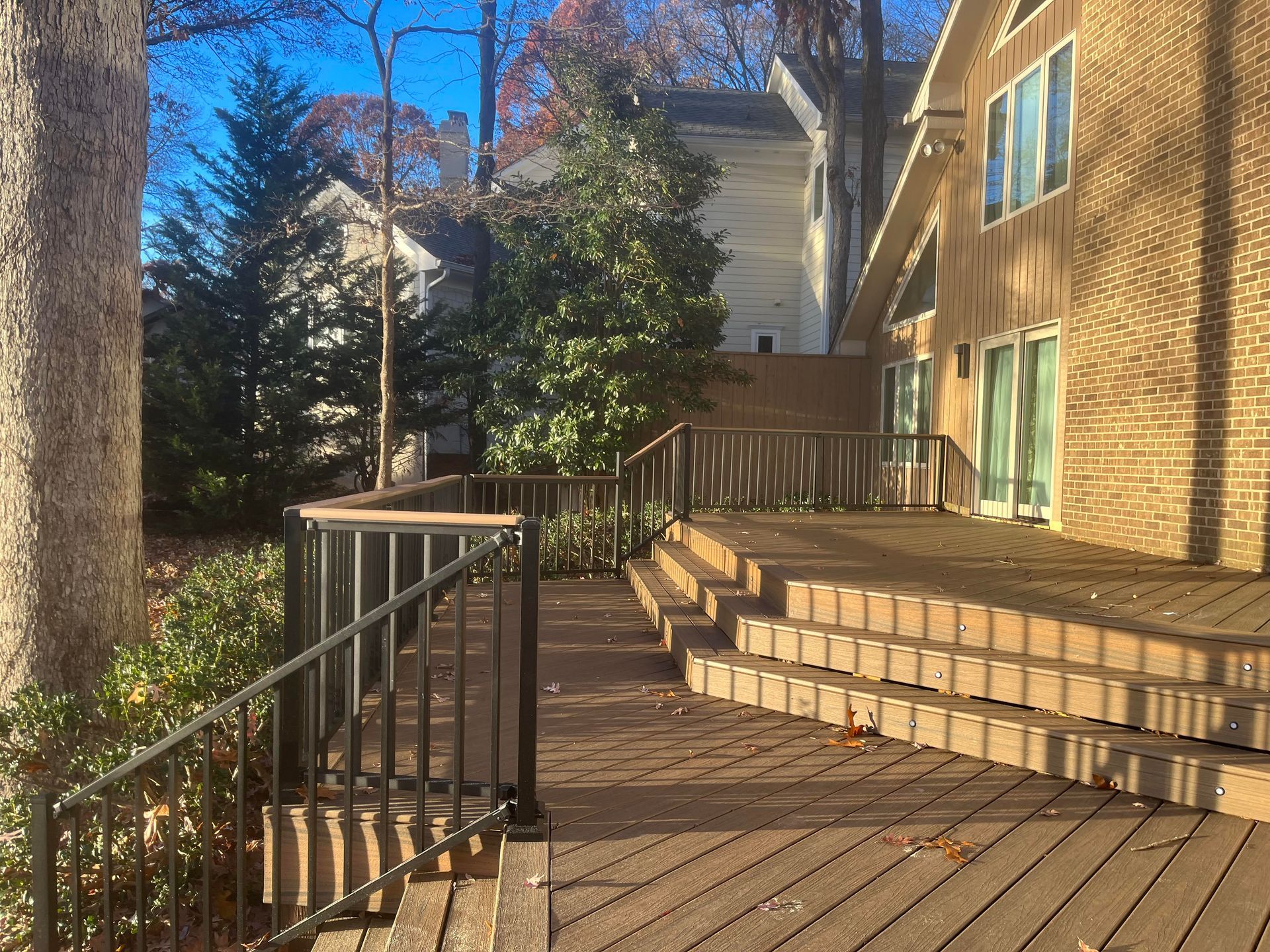 Brick apartment patio with wooden deck, black railing, steps, and trees in sunlight