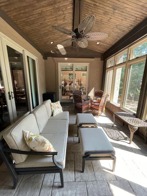 Sunroom with gray sofa, ottoman, wicker chair, and large windows letting in bright sunlight.
