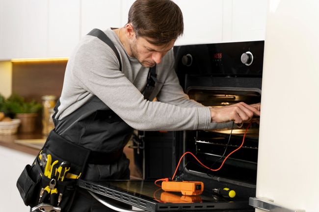 Technician repairing an oven with tools and exposed wiring in a kitchen.
