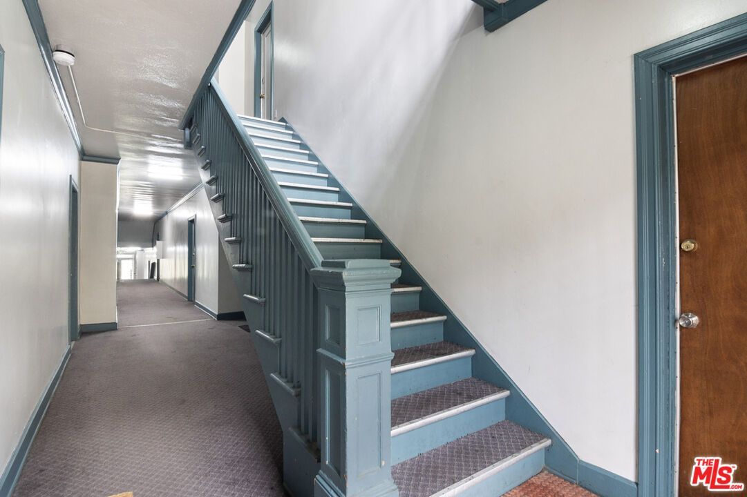 Hallway with stairs; blue banister, brown door, gray carpet, white walls.