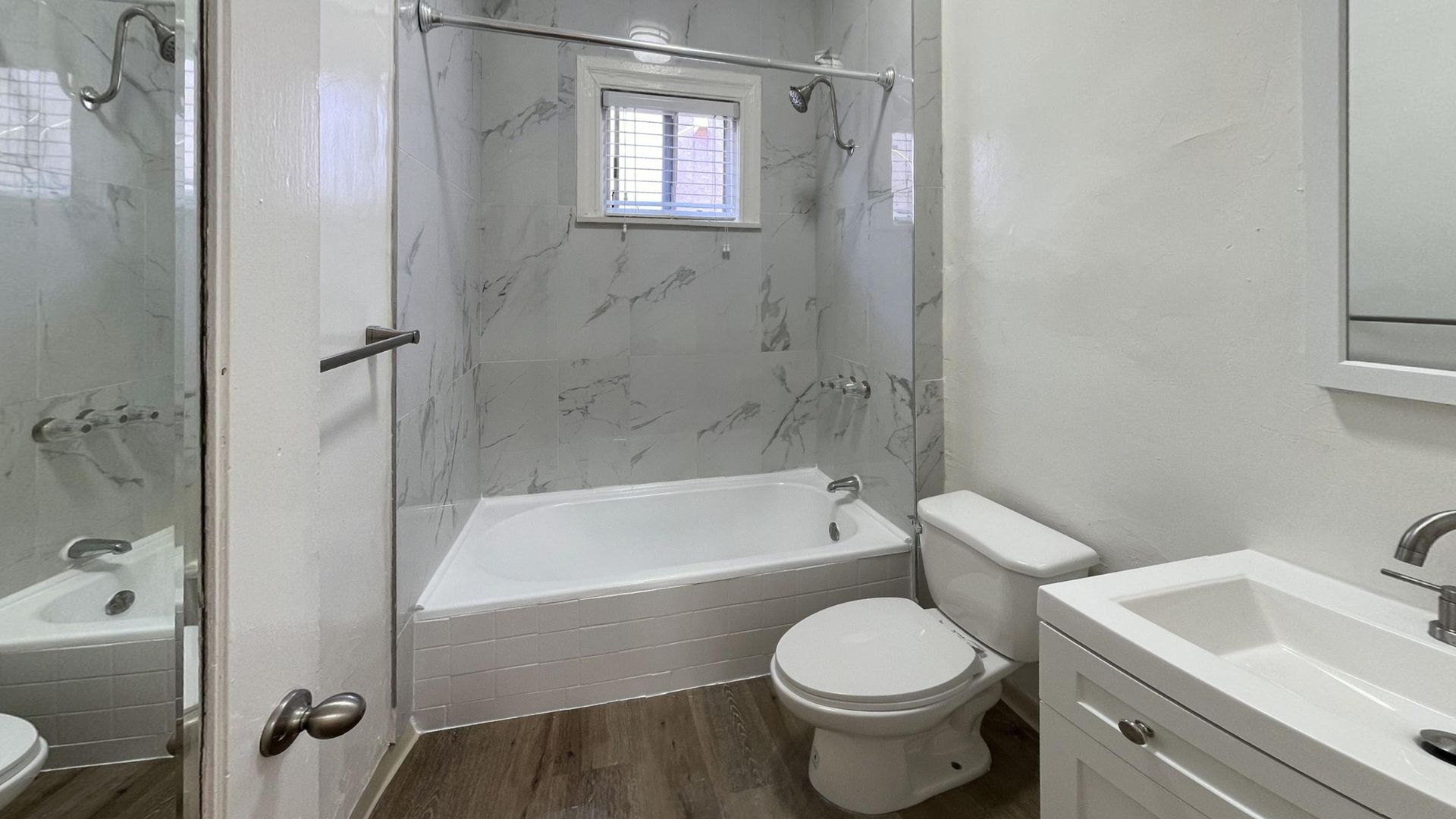 Bathroom with white walls, marble-look tiled shower, white tub, toilet, and sink, and wooden floor.