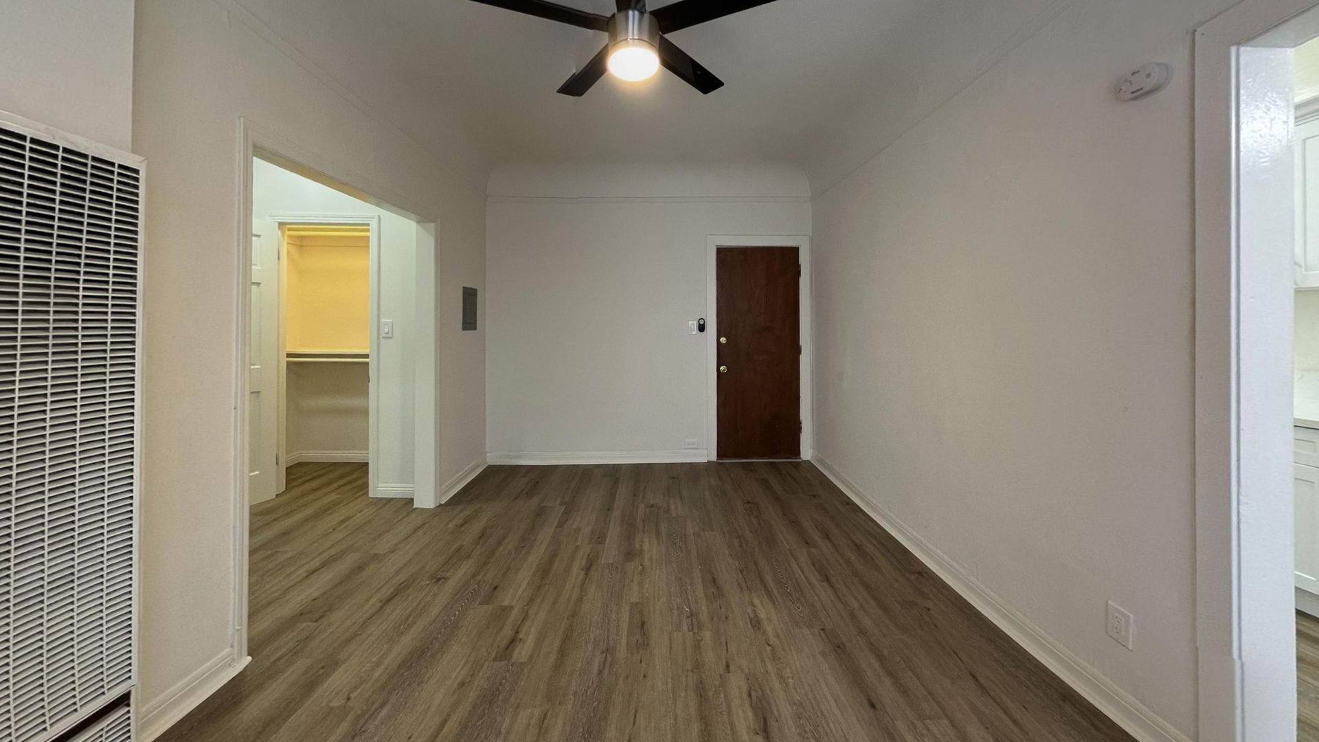 Empty room with wood-look flooring, white walls, brown door, closet entrance, and ceiling fan.