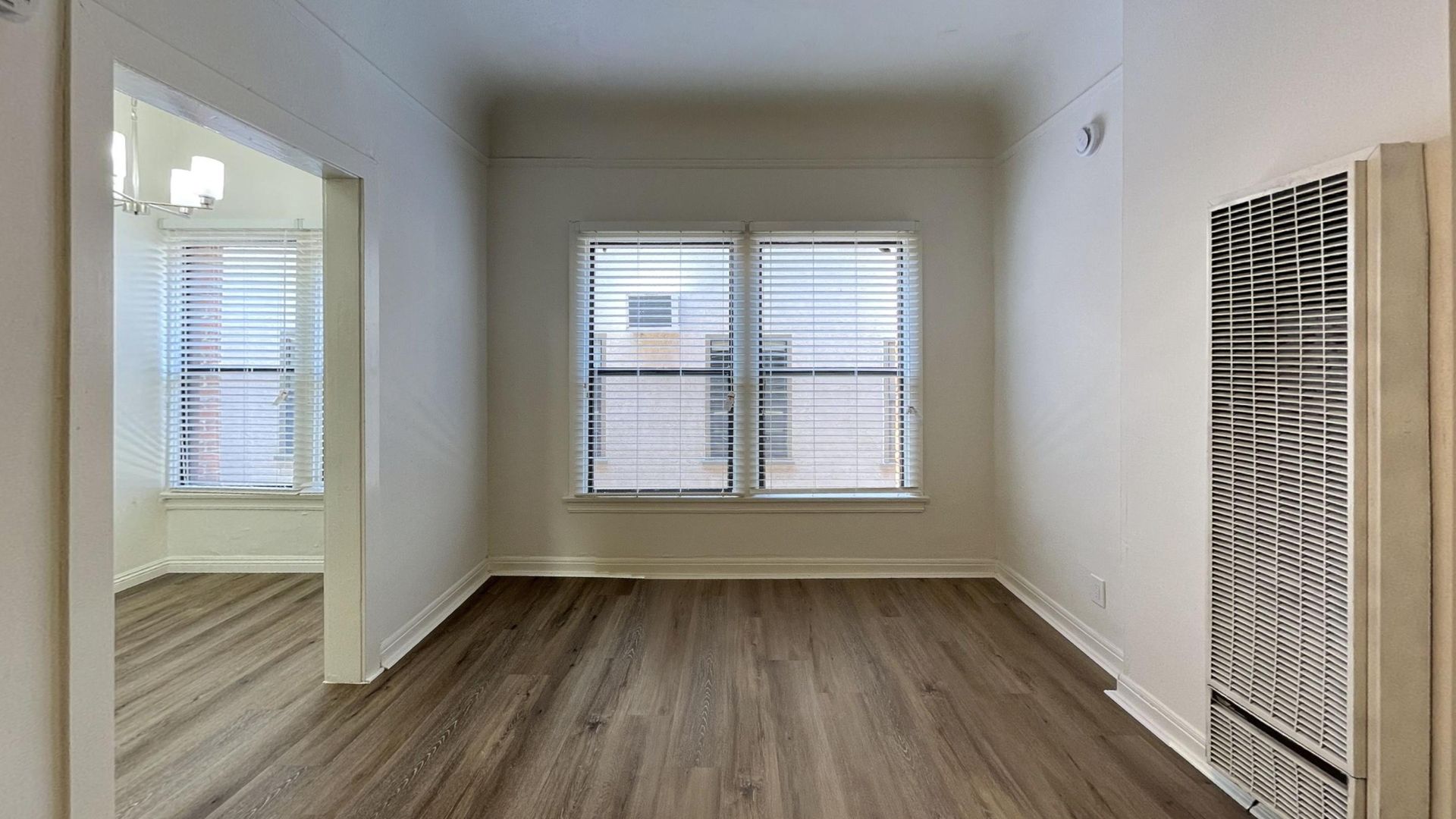 Empty room with wood-look flooring and two windows with blinds. A doorway leads to another room.