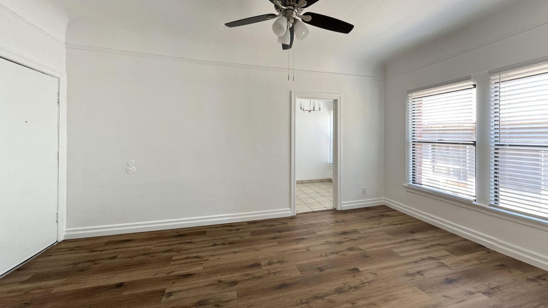 Empty room with brown wooden floor, white walls, closet, and windows with blinds. Ceiling fan present.