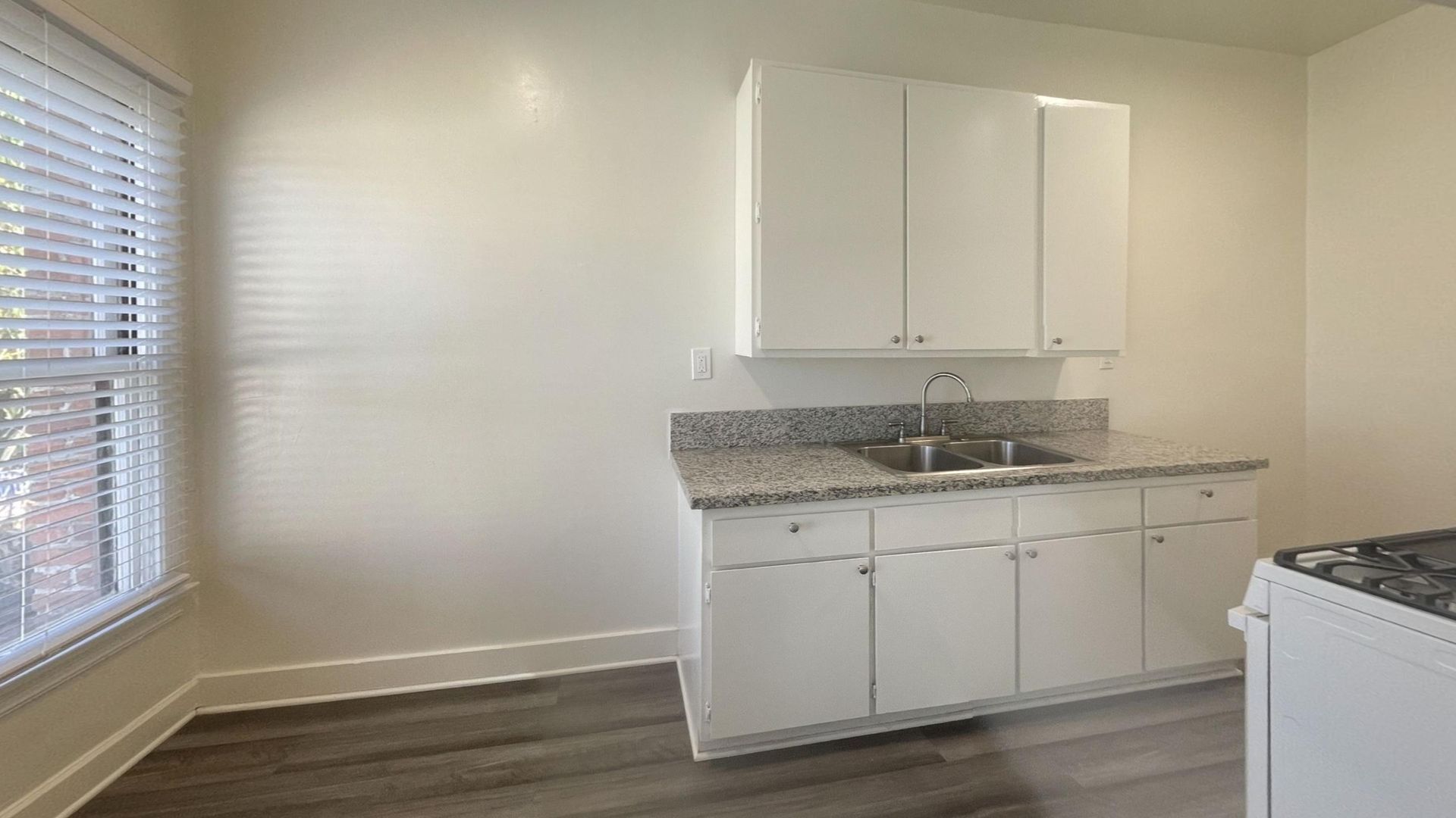 White kitchen cabinets and sink area; window with blinds; gray countertop and floor.