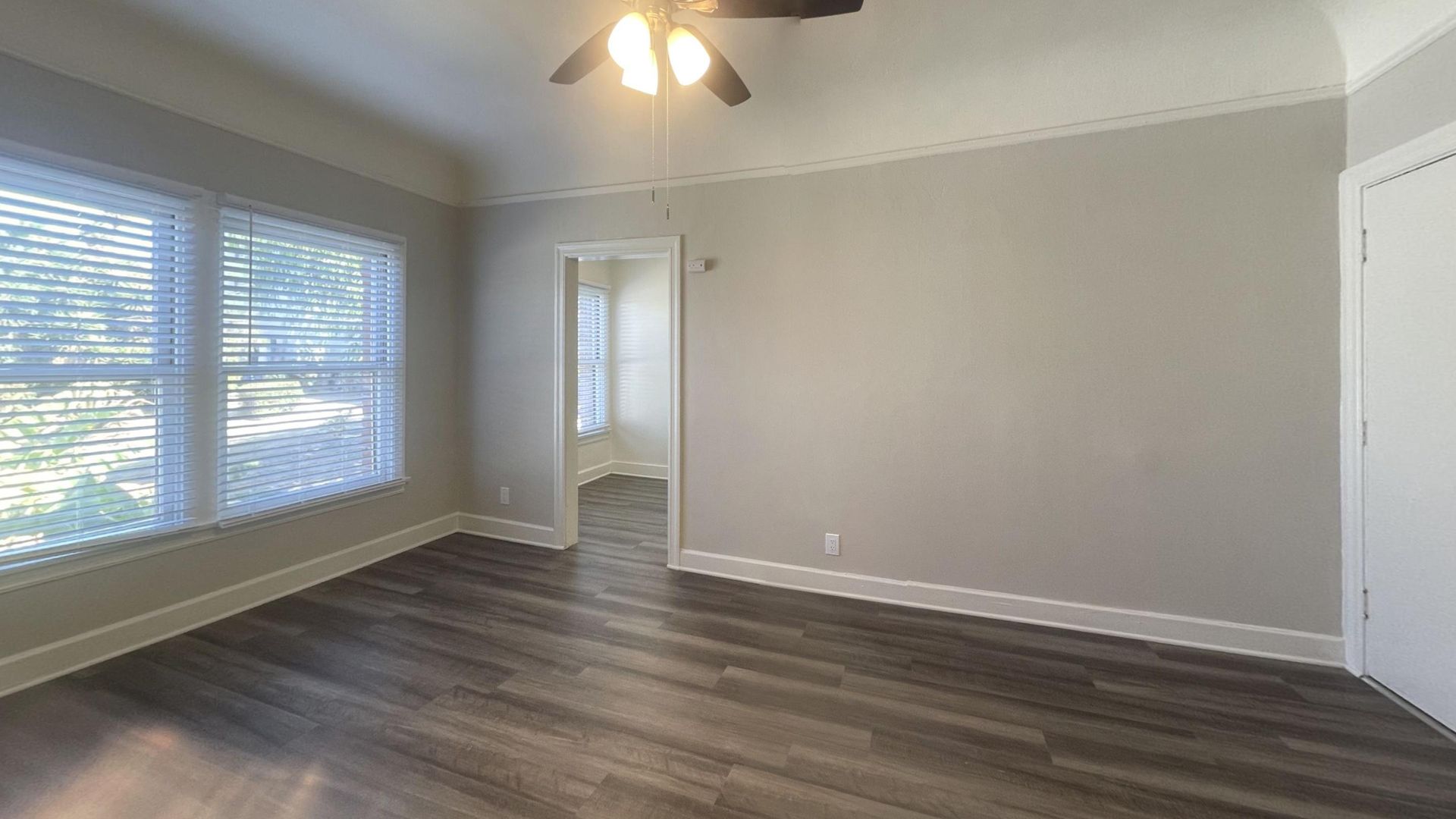 Empty room with gray walls, dark wood-look floor, windows, and a doorway. Ceiling fan and white trim.