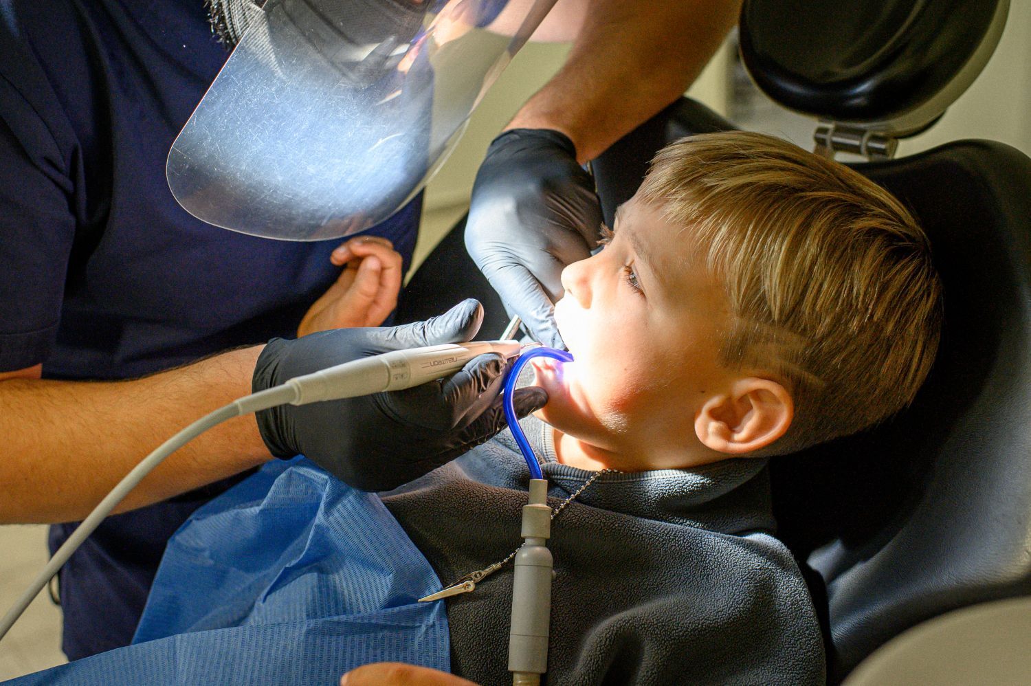 A dental professional in a face shield and black gloves cleans a patient's teeth using a dental handpiece and suction.