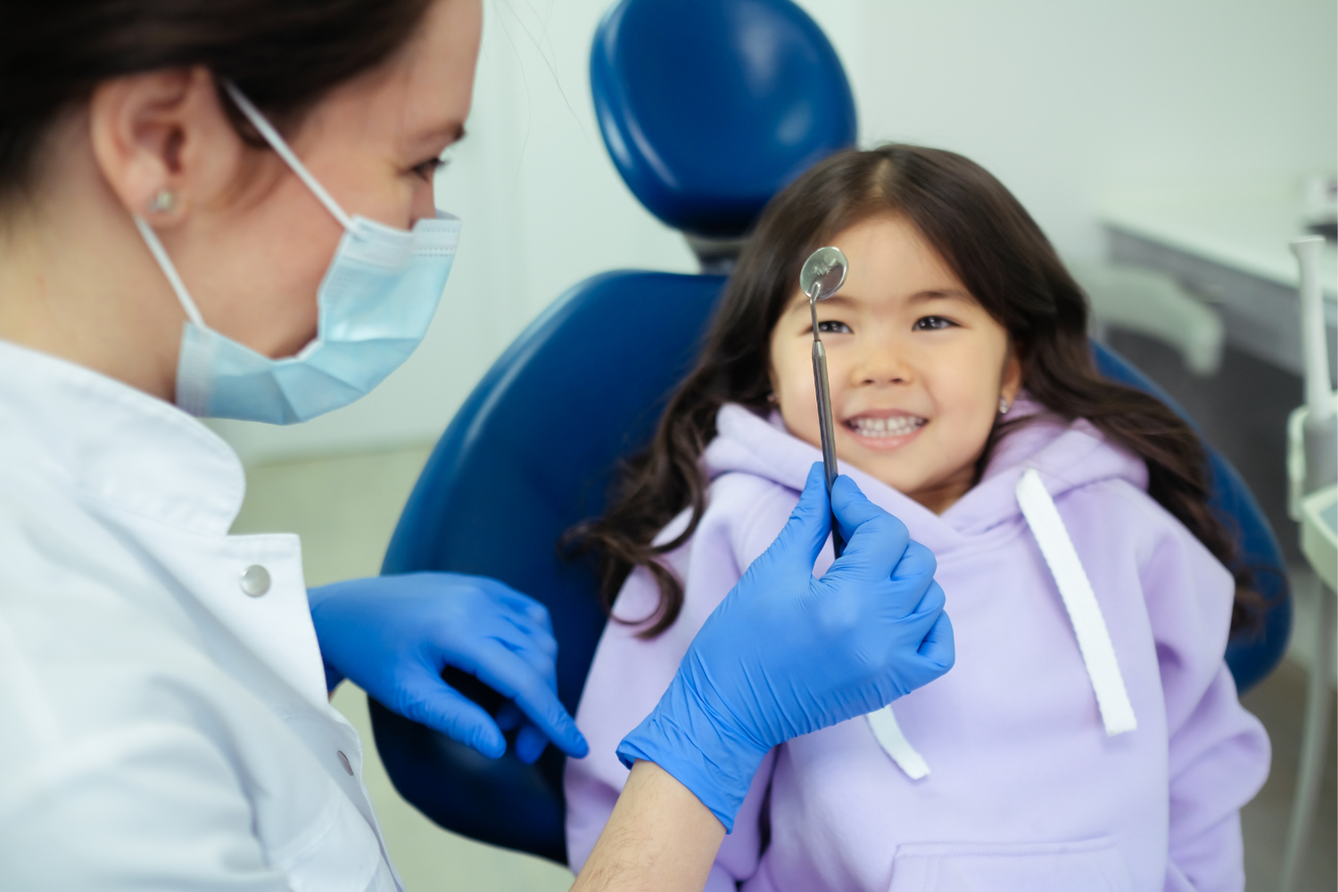 A dentist in a mask and blue gloves holds a dental mirror for a smiling patient in a lavender hoodie.