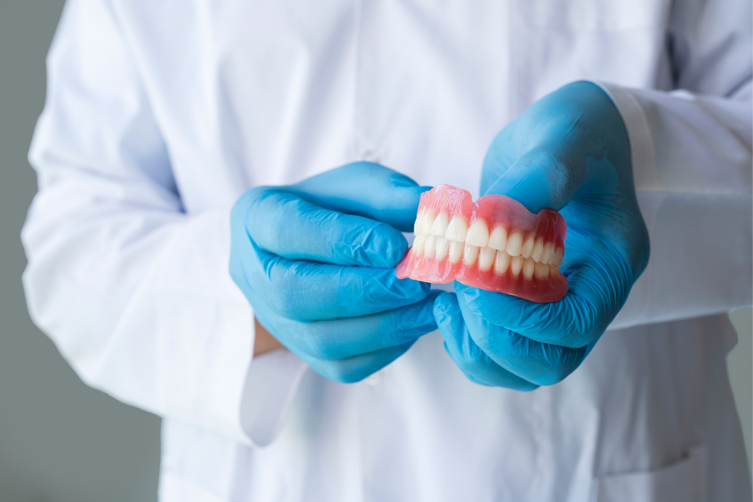 A dentist wearing a white coat and blue gloves holds a set of complete dentures.