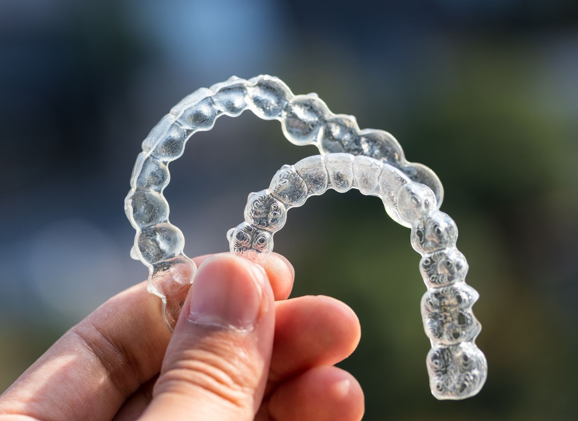A hand holds up a pair of clear plastic dental aligner trays against a blurred outdoor background.