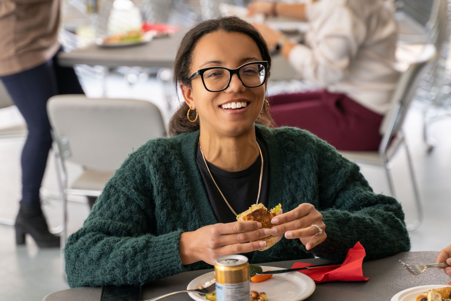 Woman smiles, holding sandwich, at table. Wearing glasses, green sweater. Eating at an indoor event.