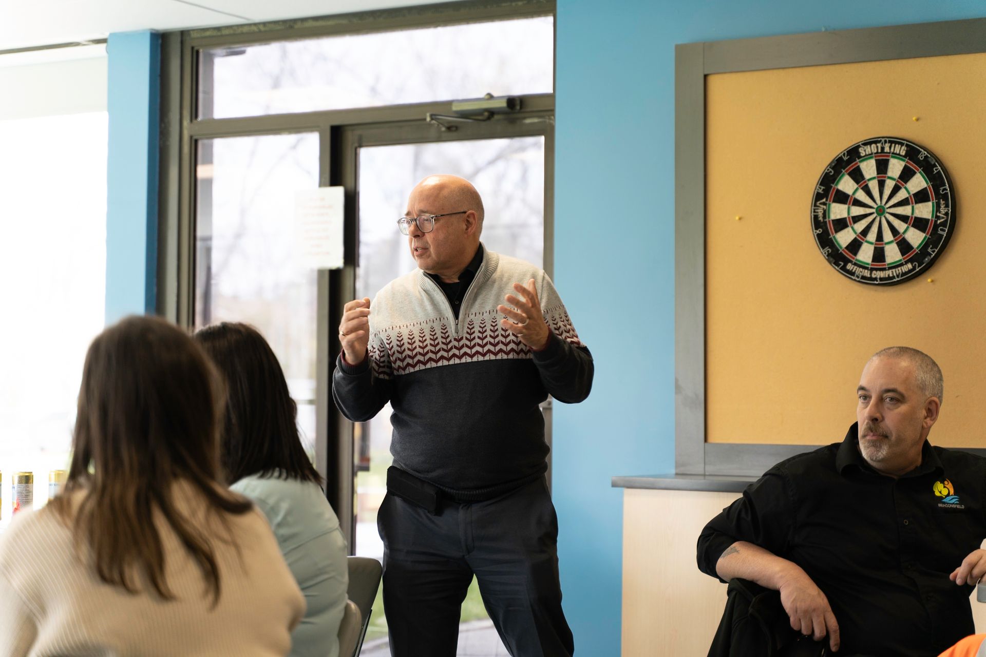 Man speaking to a group in front of a window and dartboard.