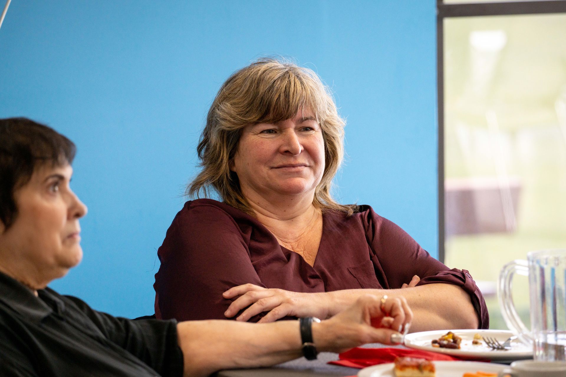 Woman in burgundy top sits at a table, looking to the right with arms crossed. Another woman is seated to the left.
