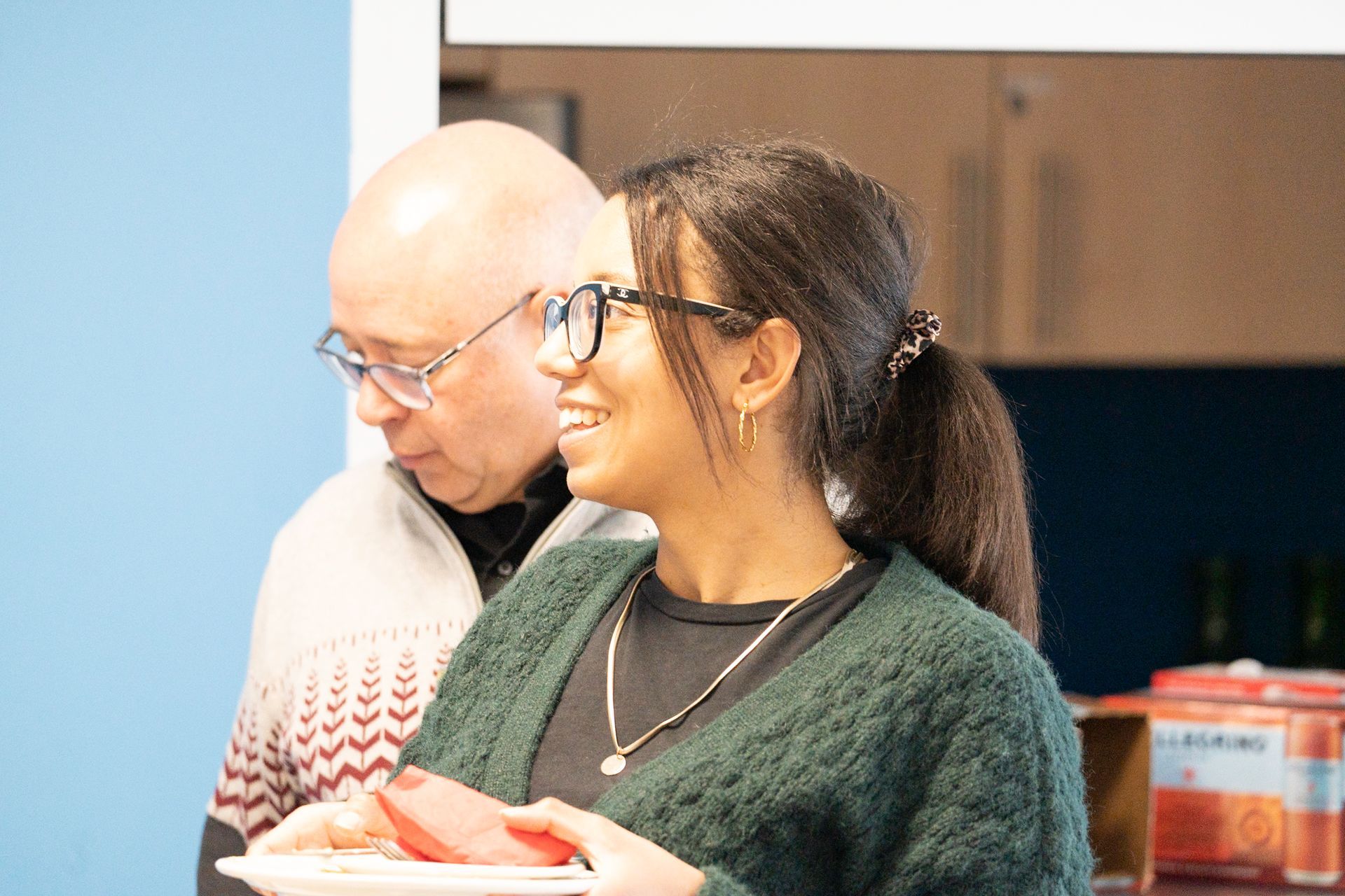 Woman smiles, holding plate with food, stands next to a man wearing glasses, in a kitchen with blue walls.