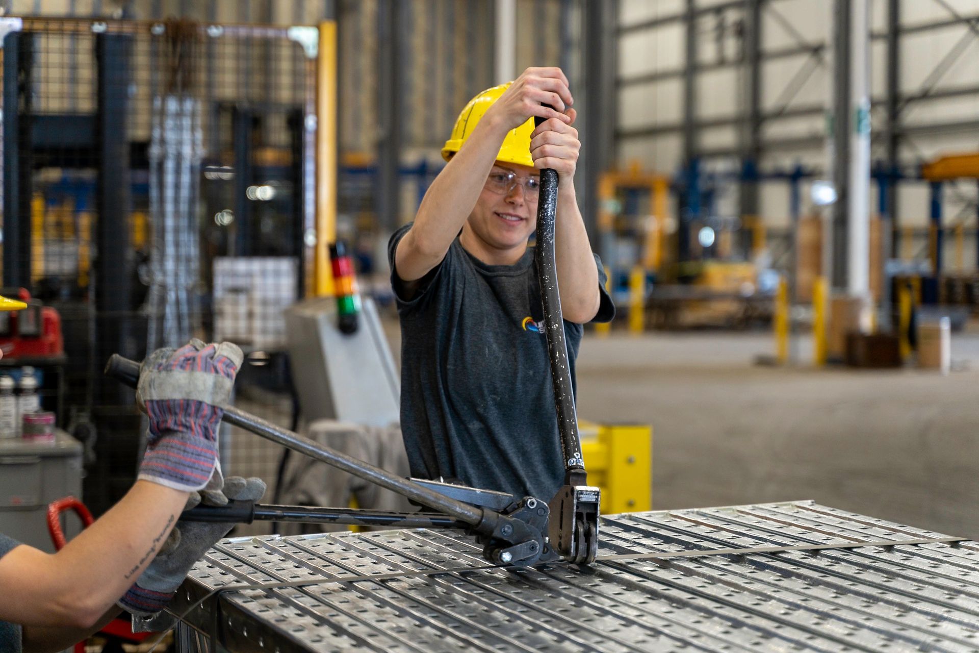 Person in a yellow hard hat cutting metal bars with a large tool in a factory setting.