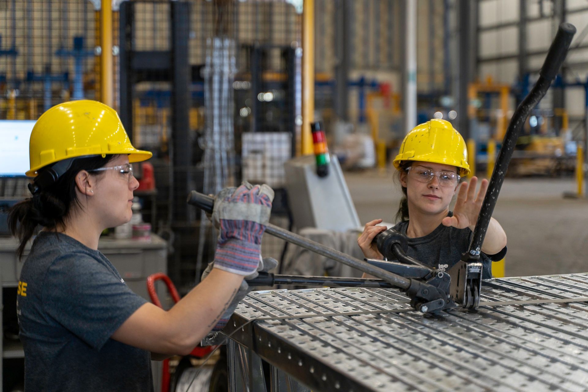 Two people wearing yellow hard hats working with metal in a factory.
