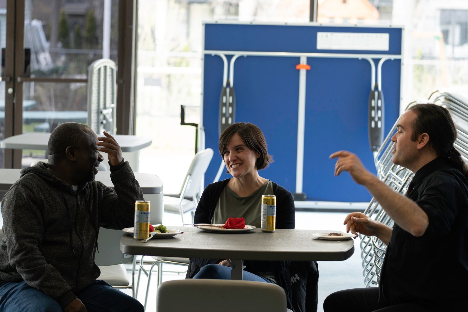 Three people seated at a table, conversing.  A ping pong table is visible in the background. Natural light floods the room.