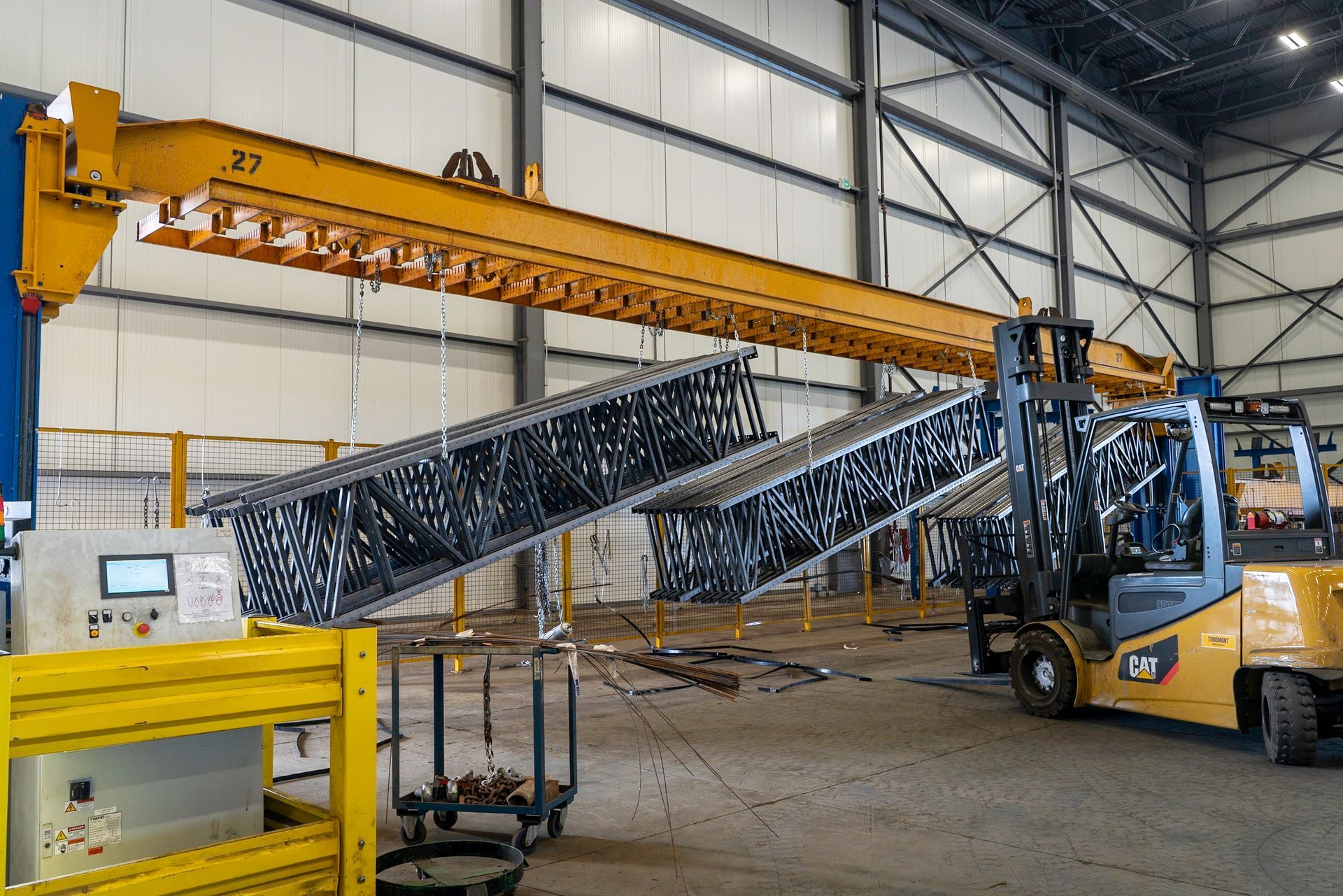 Yellow overhead crane lifting metal structures in a factory, with a forklift nearby.