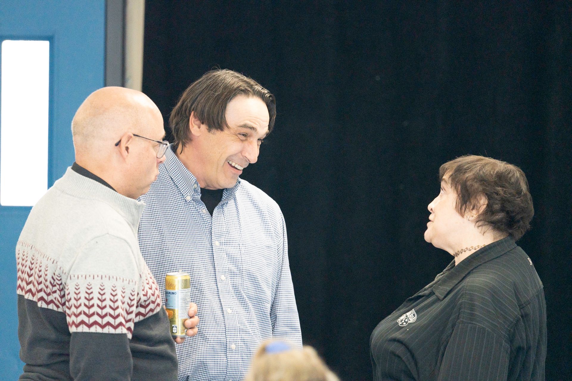 Three people conversing indoors; man with can smiles, another man and woman look on. Blue door in background.