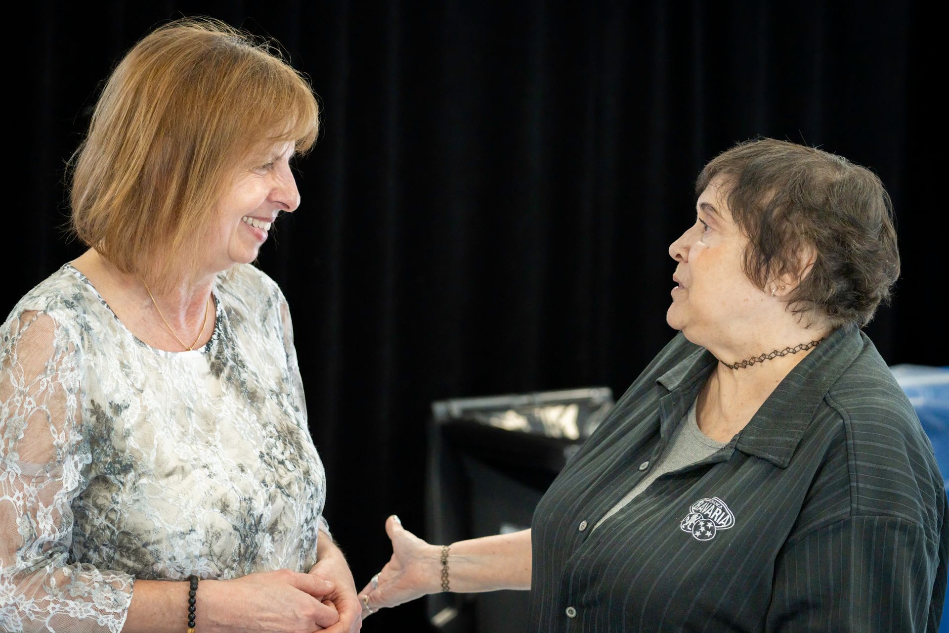 Two women talking, one gesturing, set against a dark background. One wears lace, the other a dark shirt.