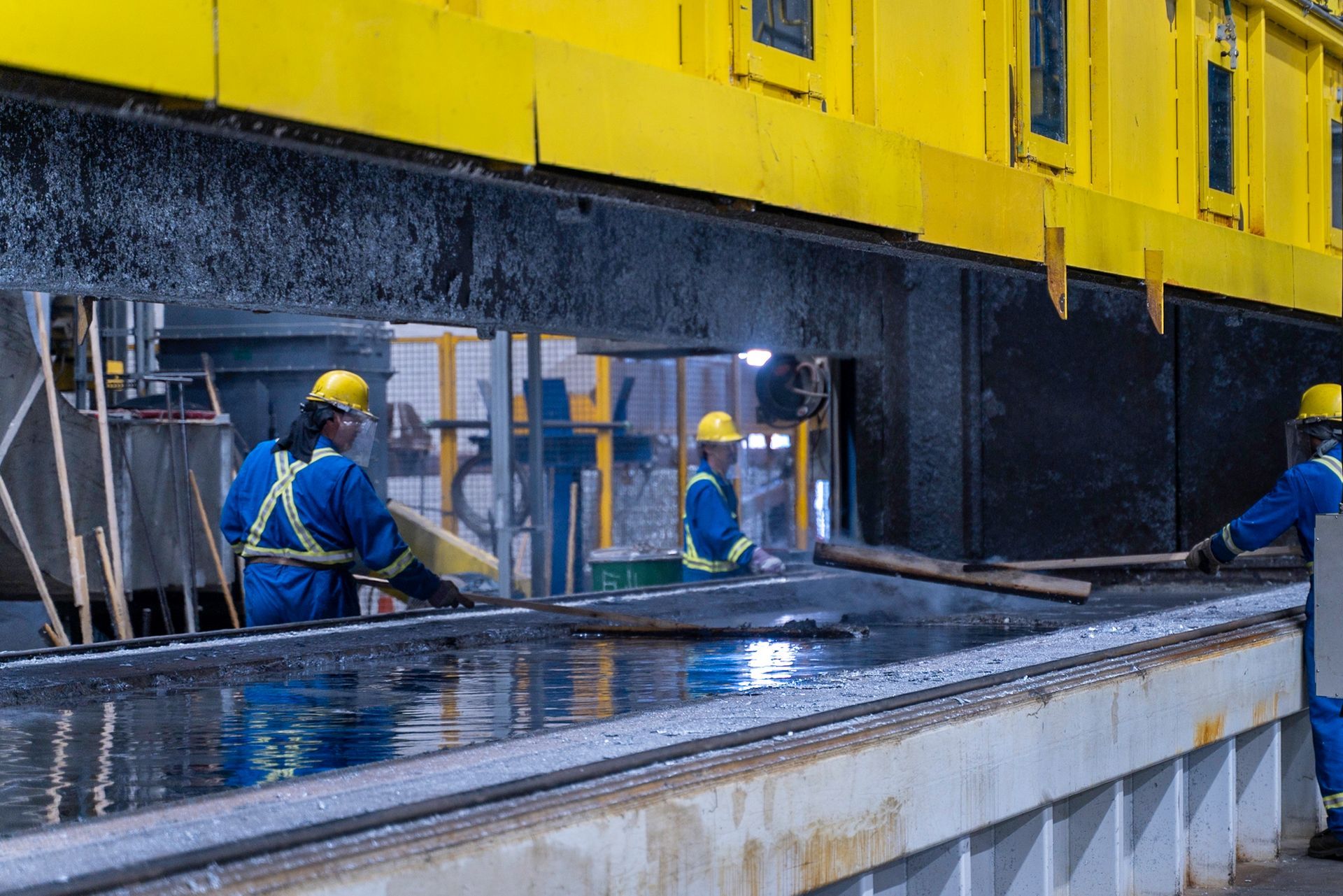 Workers in blue coveralls and yellow helmets tending to a metal processing machine.
