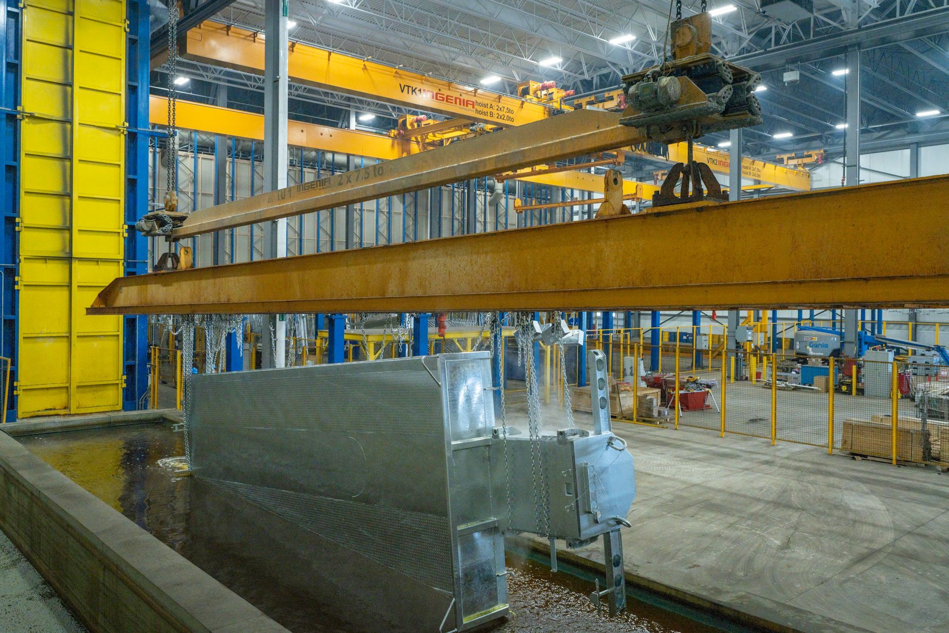 Overhead crane lowering metal object into industrial tank; yellow beams, concrete floor.
