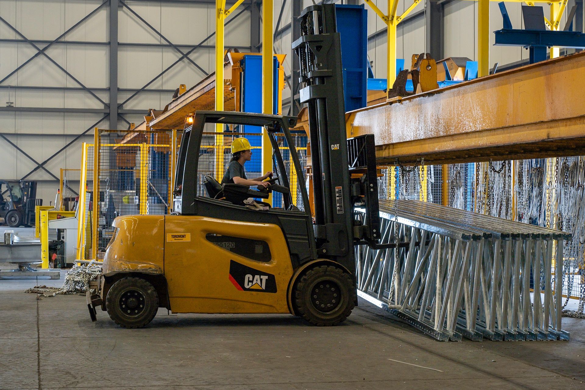 Yellow forklift in a factory transporting a metal framework. A worker wearing a yellow hard hat is driving.