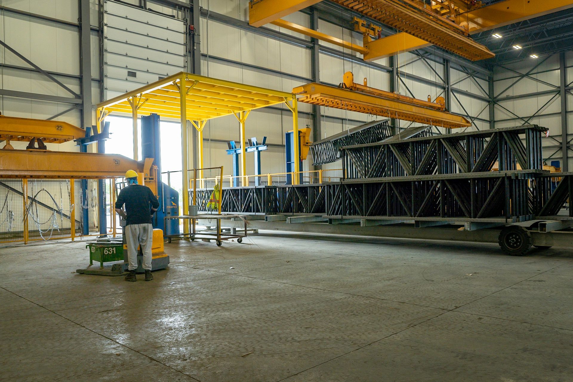 Factory interior with worker near a large metal structure, overhead cranes, and yellow scaffolding.