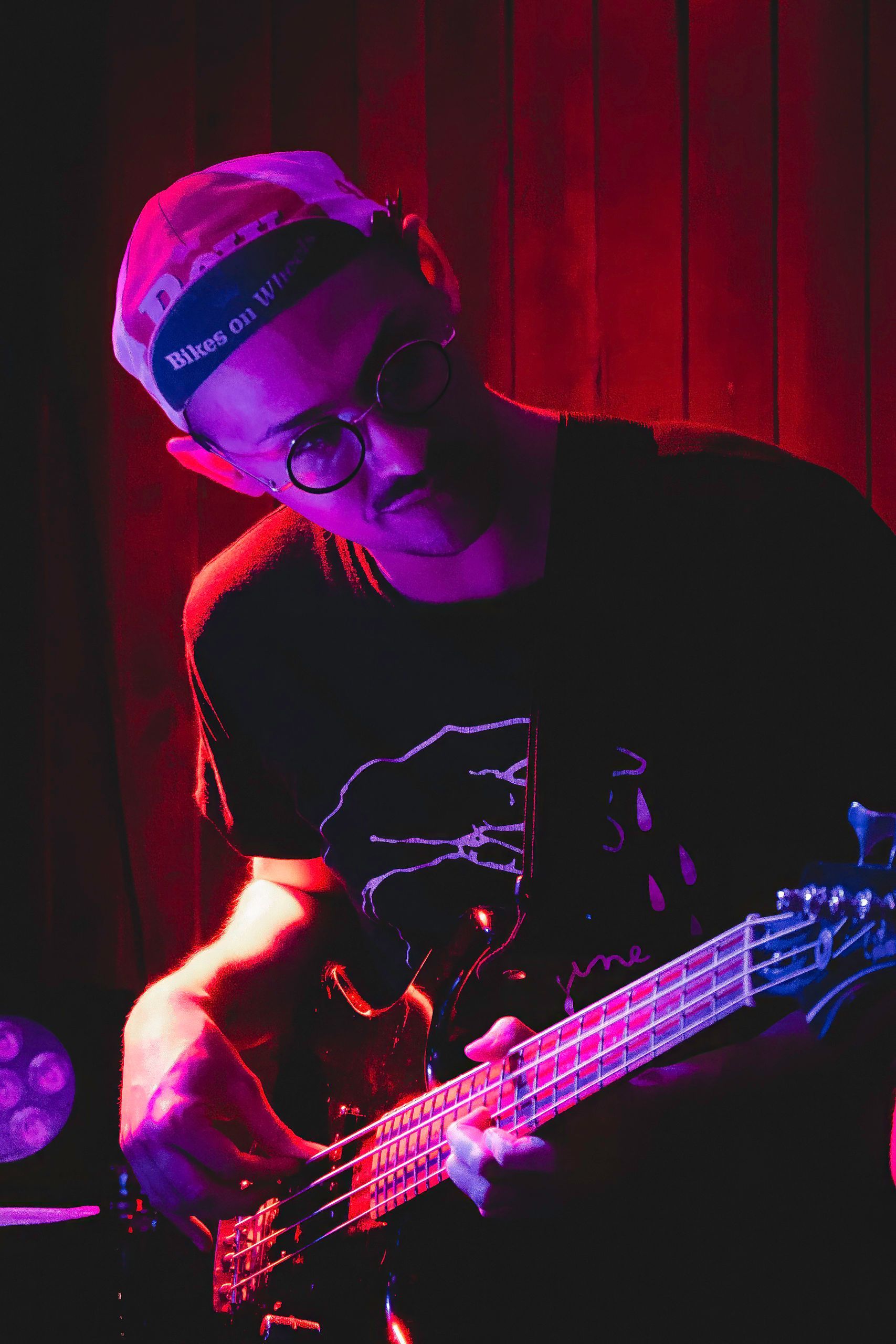 Person playing bass guitar under red and blue stage lights, wearing a bandanna.