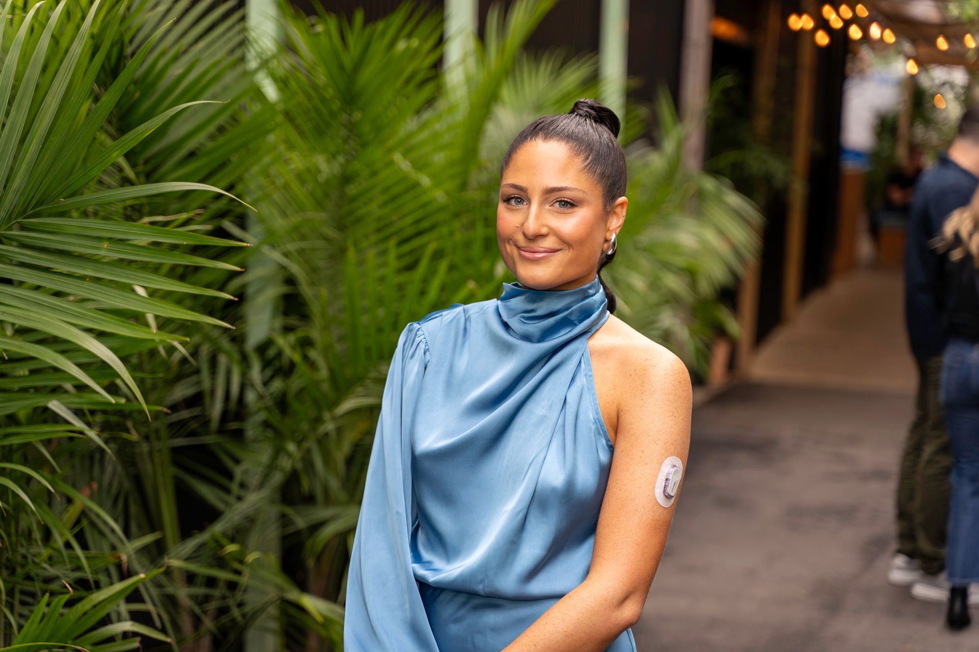 Woman in blue dress smiles outdoors, arm with medical sensor.