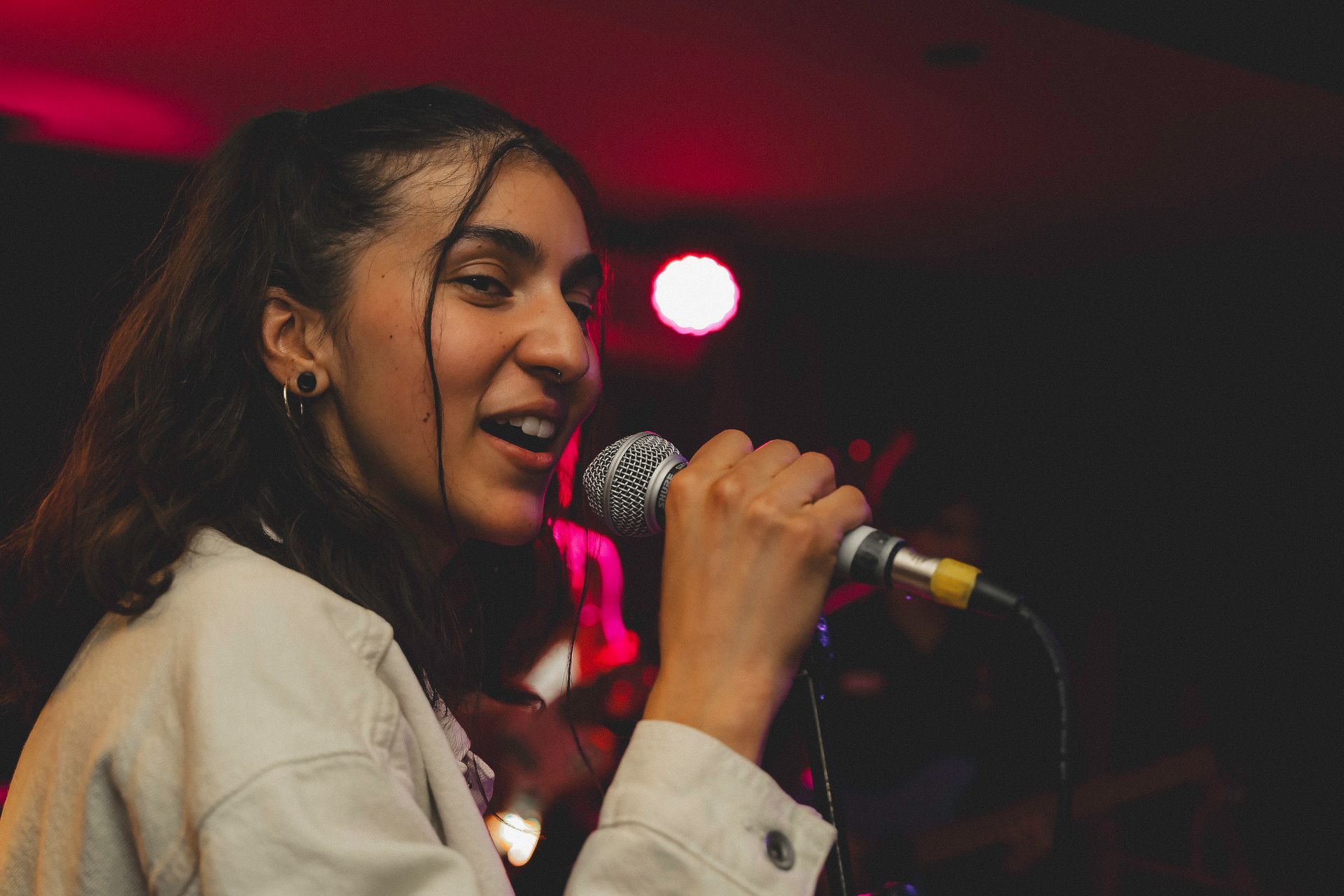 Woman singing into a microphone onstage under red lights. She wears a light jacket and has a joyful expression.