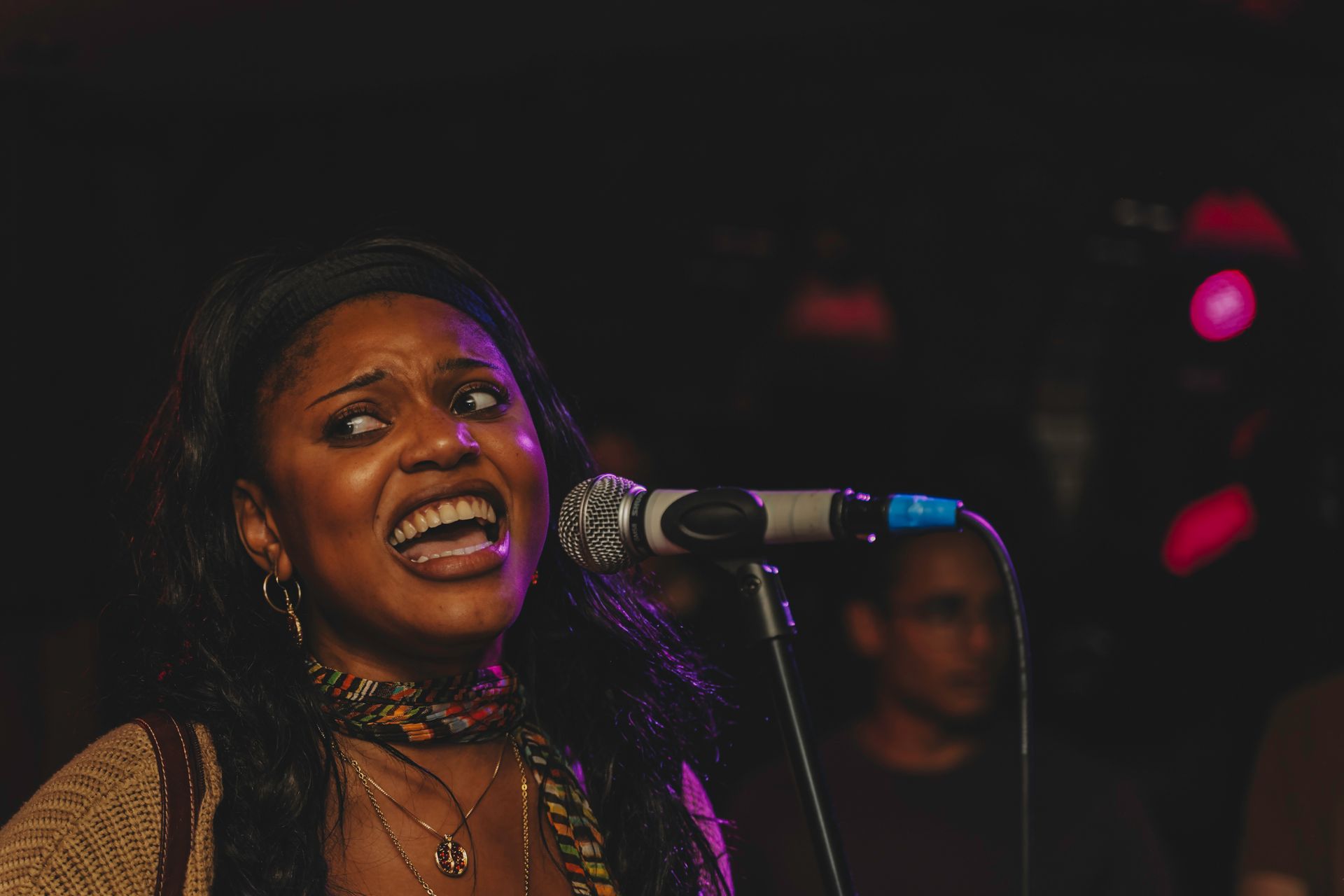 Woman singing passionately into a microphone on stage, with dark hair, a colorful scarf, and an expressive face.