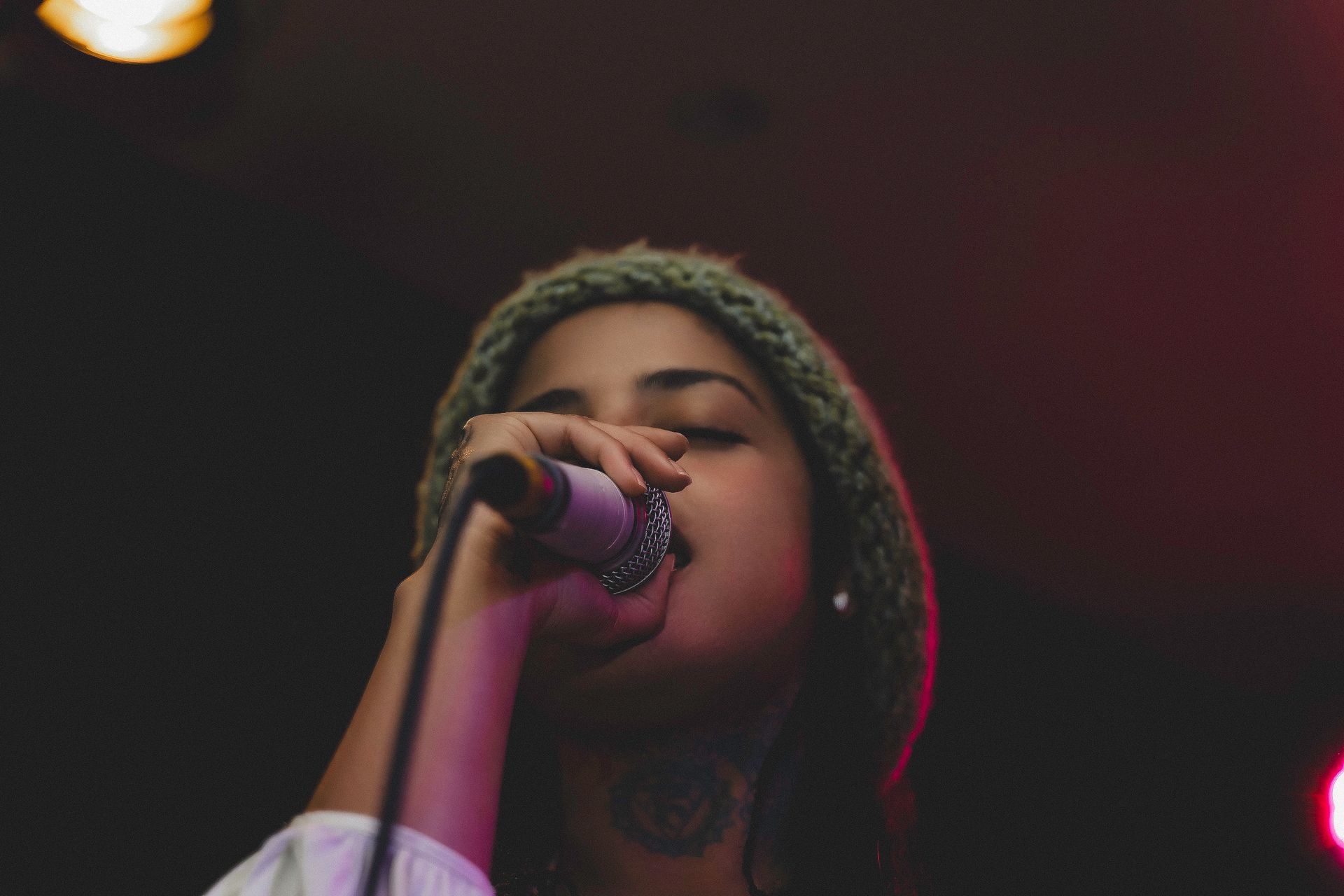 Woman singing into a microphone, wearing a beanie. Stage lighting illuminates the scene.
