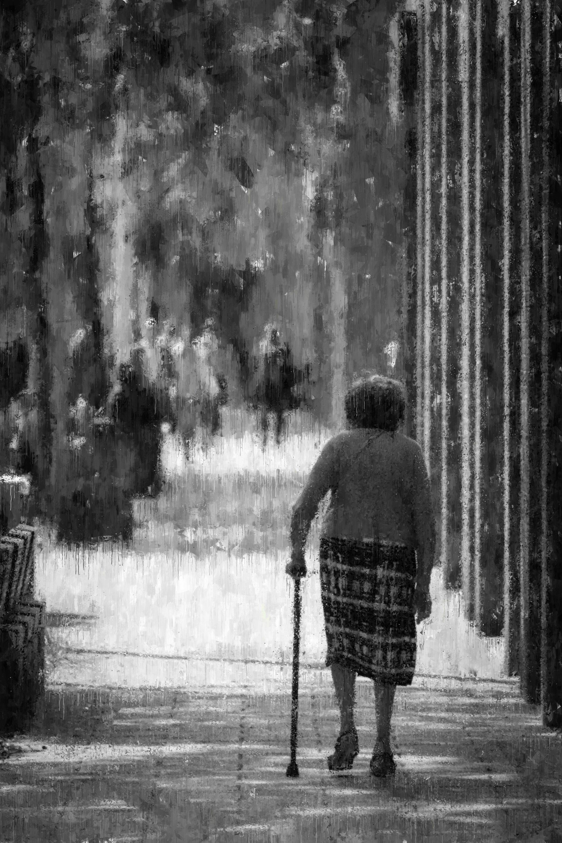 Elderly person walks with a cane along a path, trees and structures in the background. Black and white.