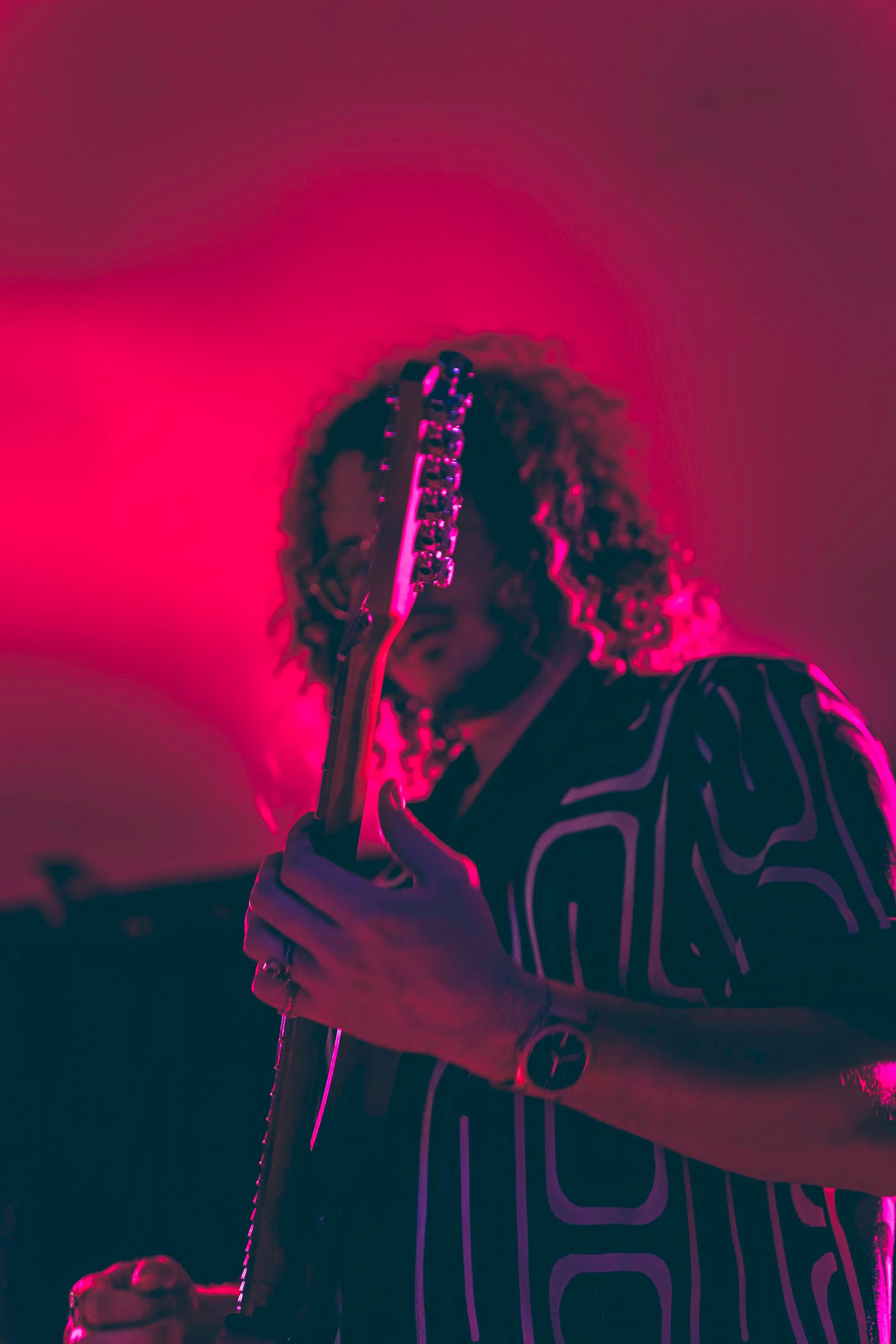 A person with curly hair playing a guitar under pink stage lights.