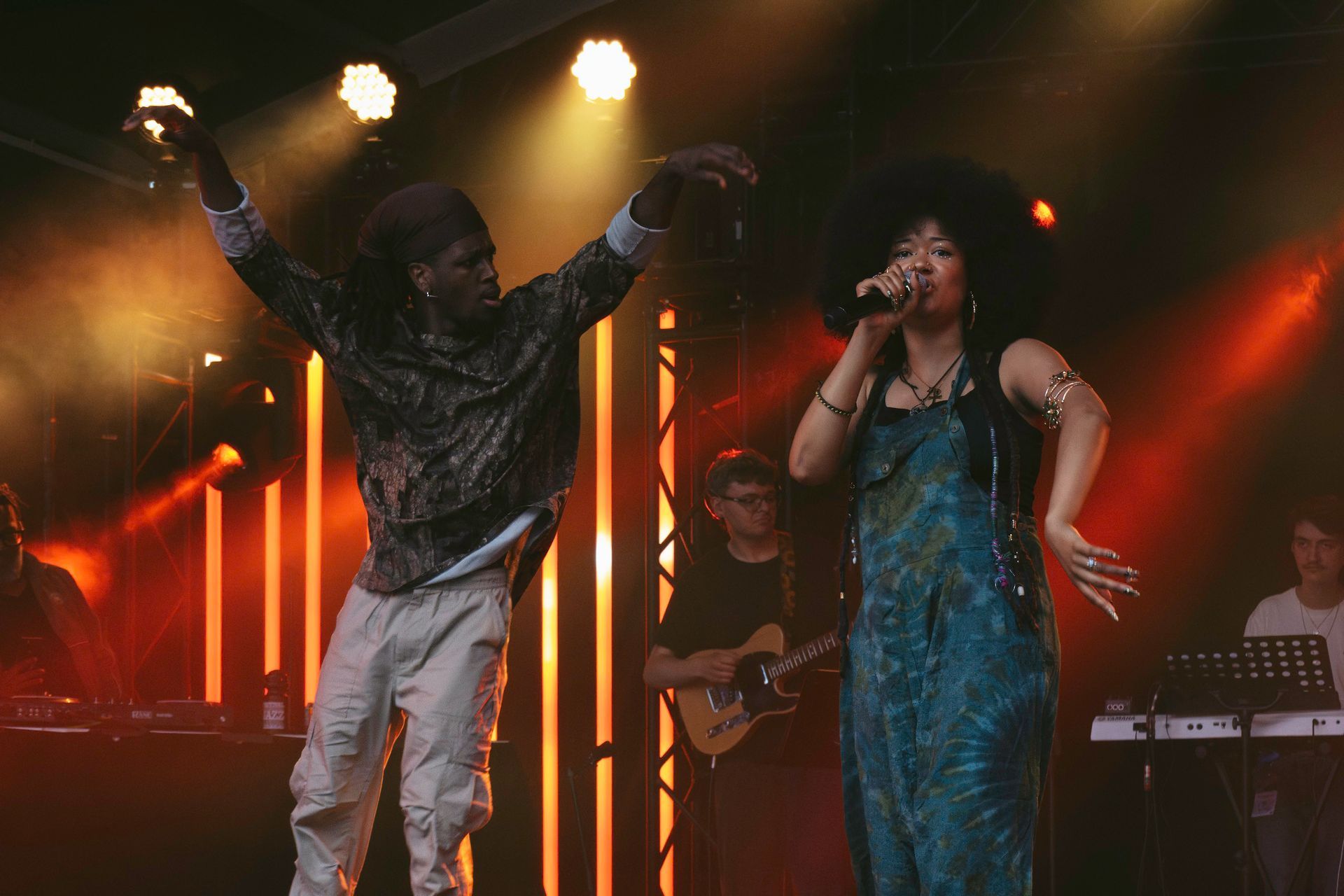 A singer with a microphone and a dancer on stage with a band, lit by stage lights.