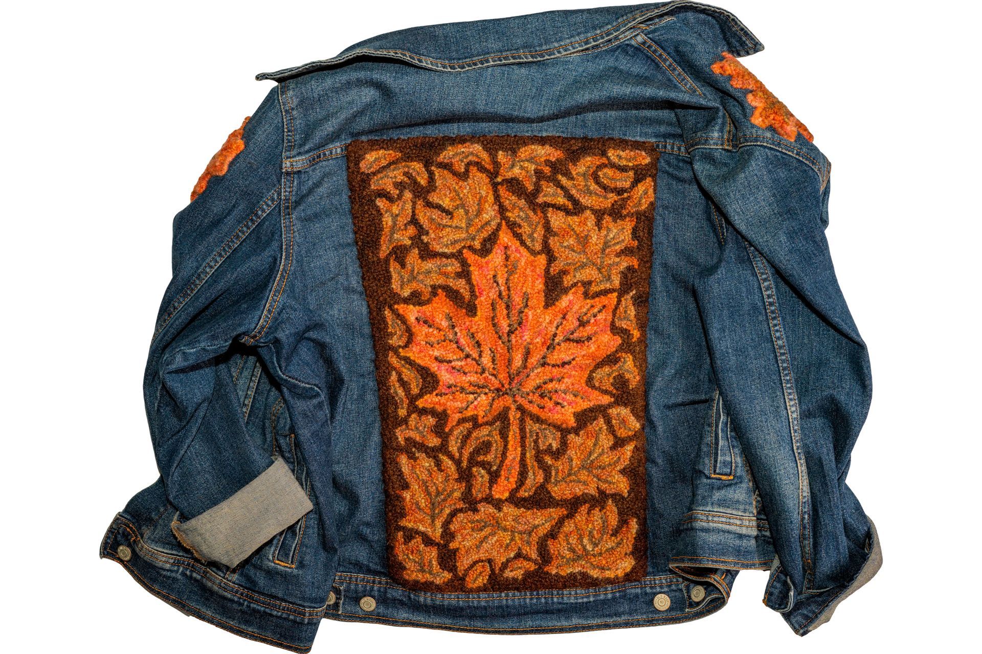 Blue denim jacket with a large embroidered autumn leaf design on the back in shades of brown and orange.