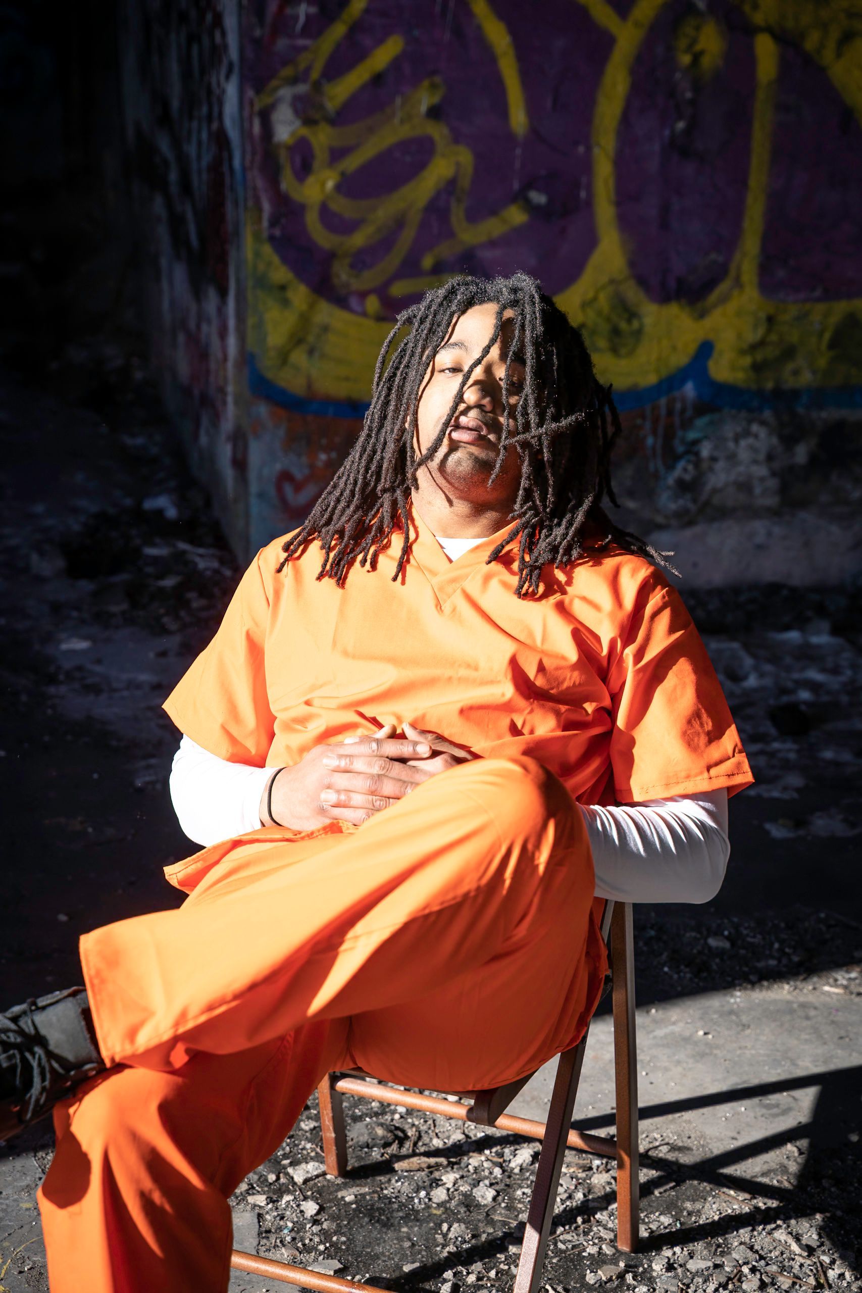Person in orange jumpsuit sits on a chair, eyes closed. Graffiti background, sunny.