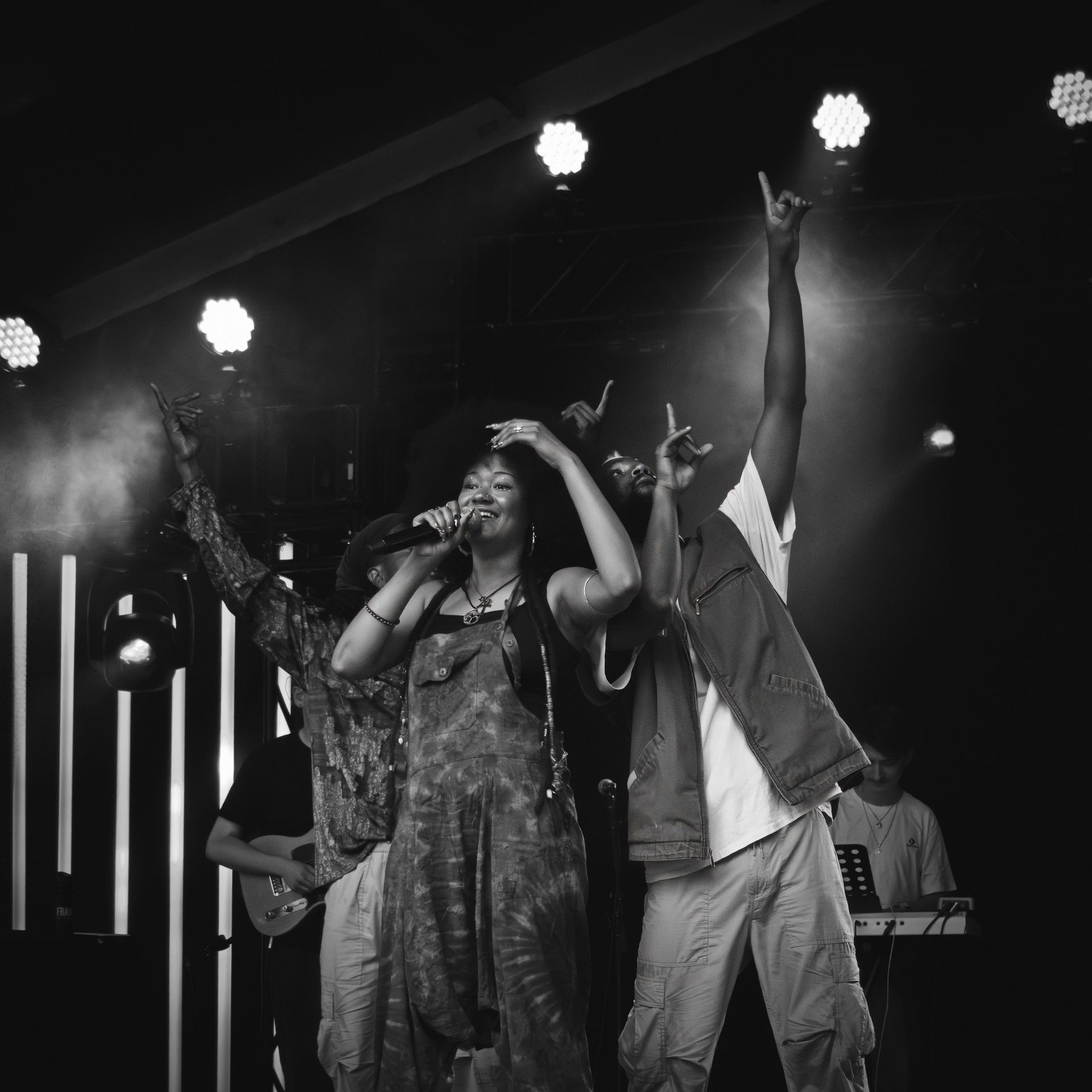 Band on stage performing, raising hands, in front of lights, microphone, and instruments. Black and white.