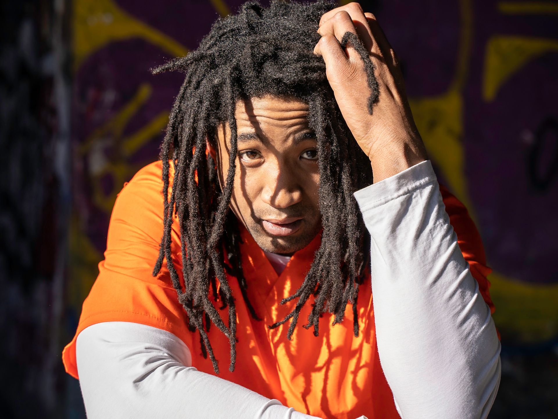 Man in orange shirt with dreadlocks, hand in hair, looking at camera. Graffiti background.