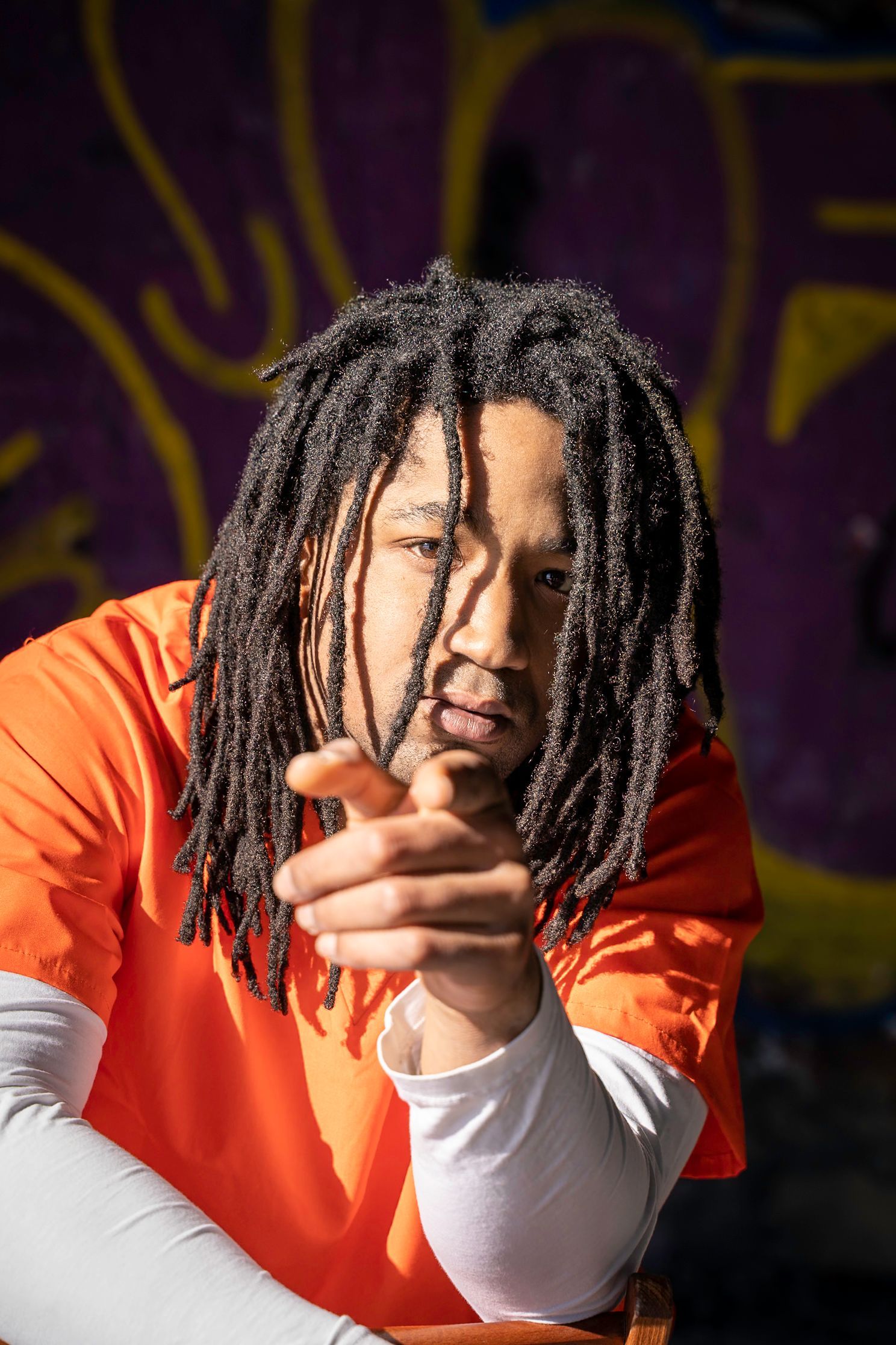 Man with dreadlocks, orange shirt, pointing. Graffiti background.