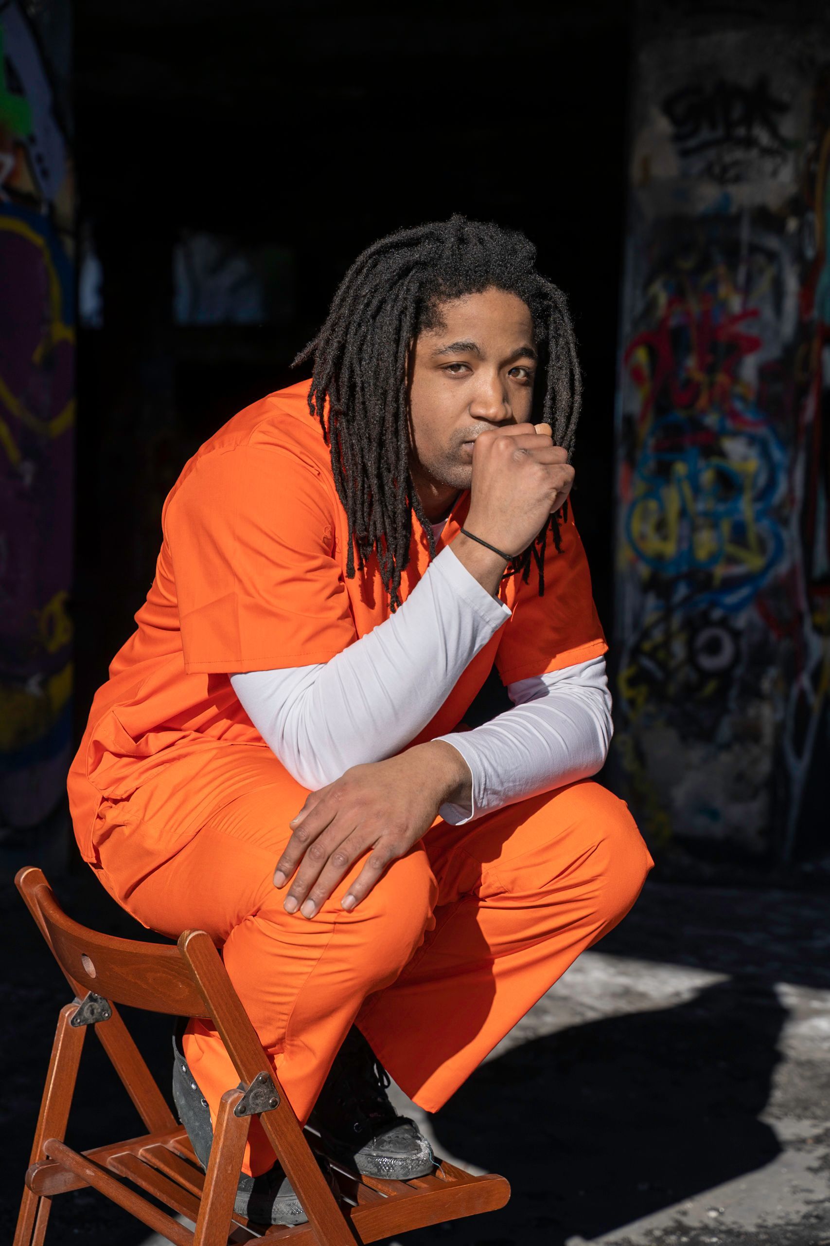 Man in orange jumpsuit squats on wooden chair, chin resting on hand, in front of graffiti-covered pillars.