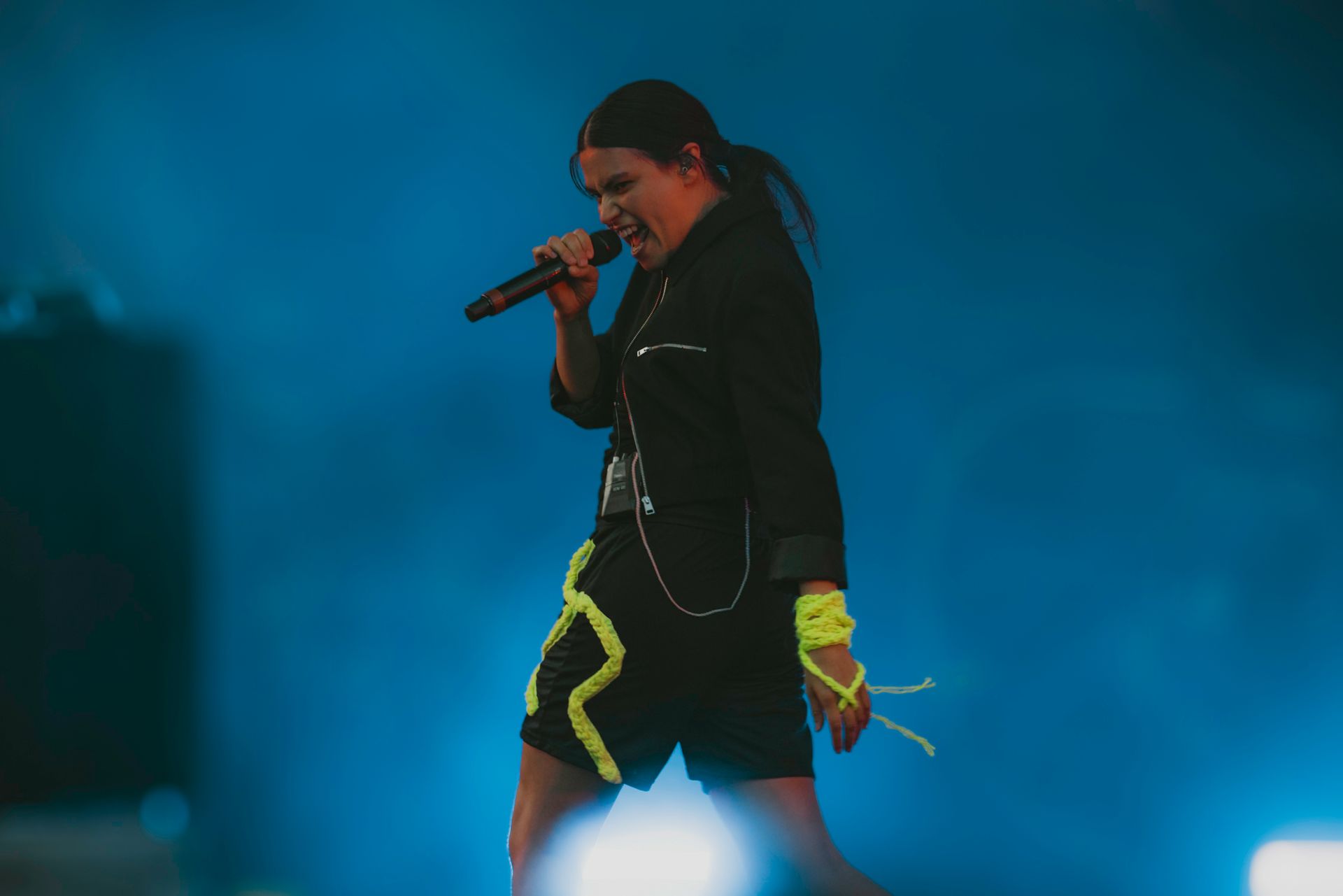 Singer on stage, wearing a black outfit with neon green accents, holding a microphone. Blue background.