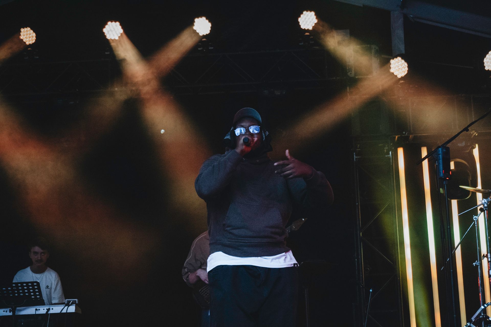 Person performing on stage, dark hoodie and sunglasses, gesturing, stage lights.