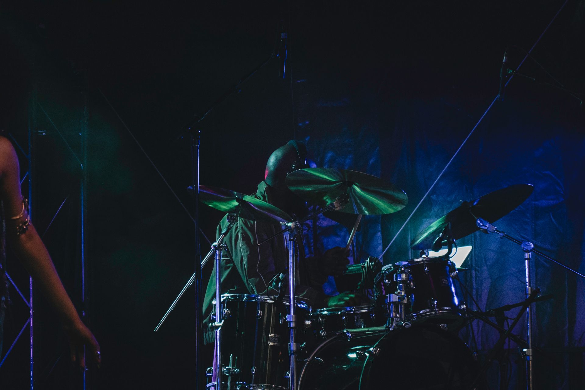 Drummer playing a drum kit on stage under green and blue stage lights.