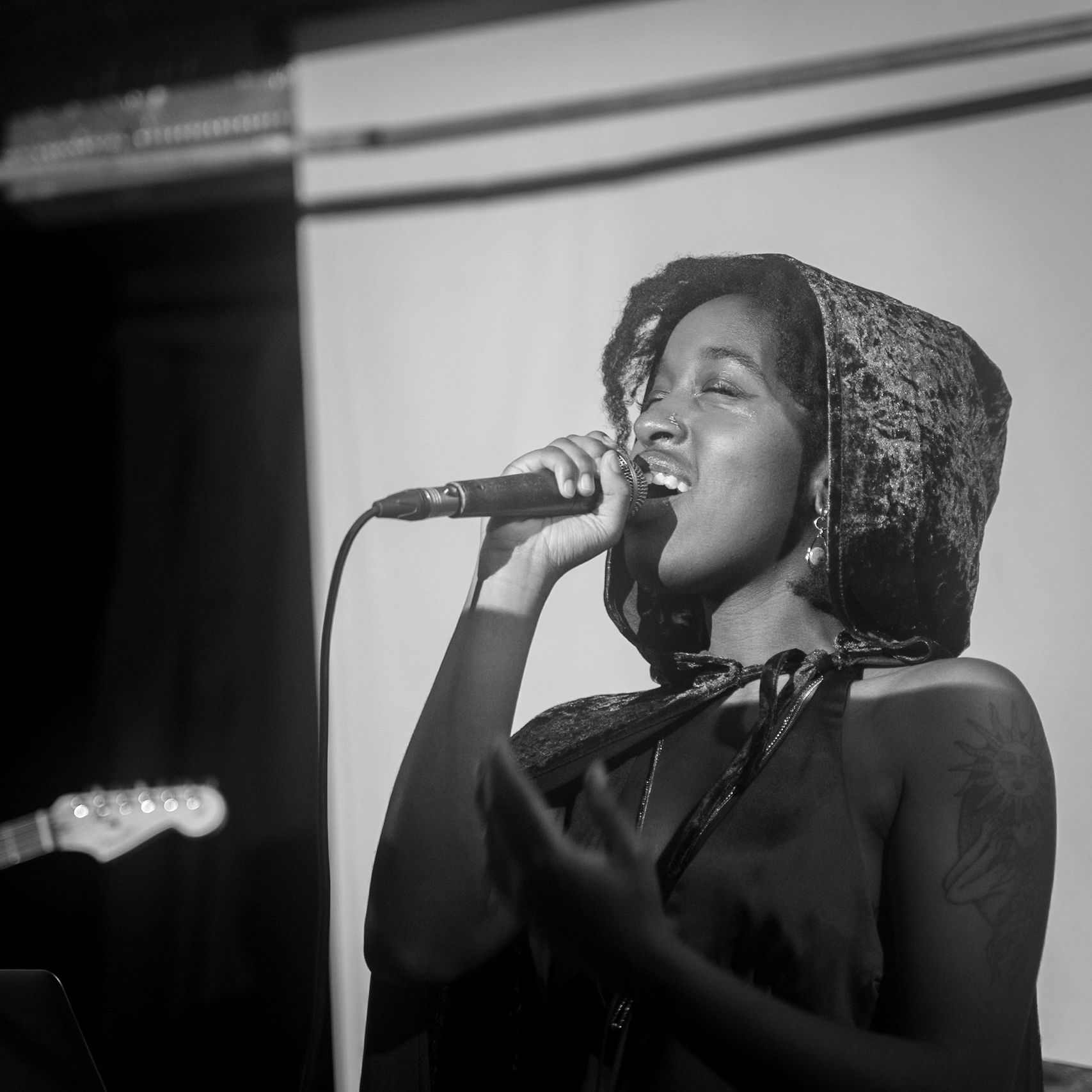 Woman singing into a microphone, wearing hooded garment. Indoor performance setting, guitar visible on the left.
