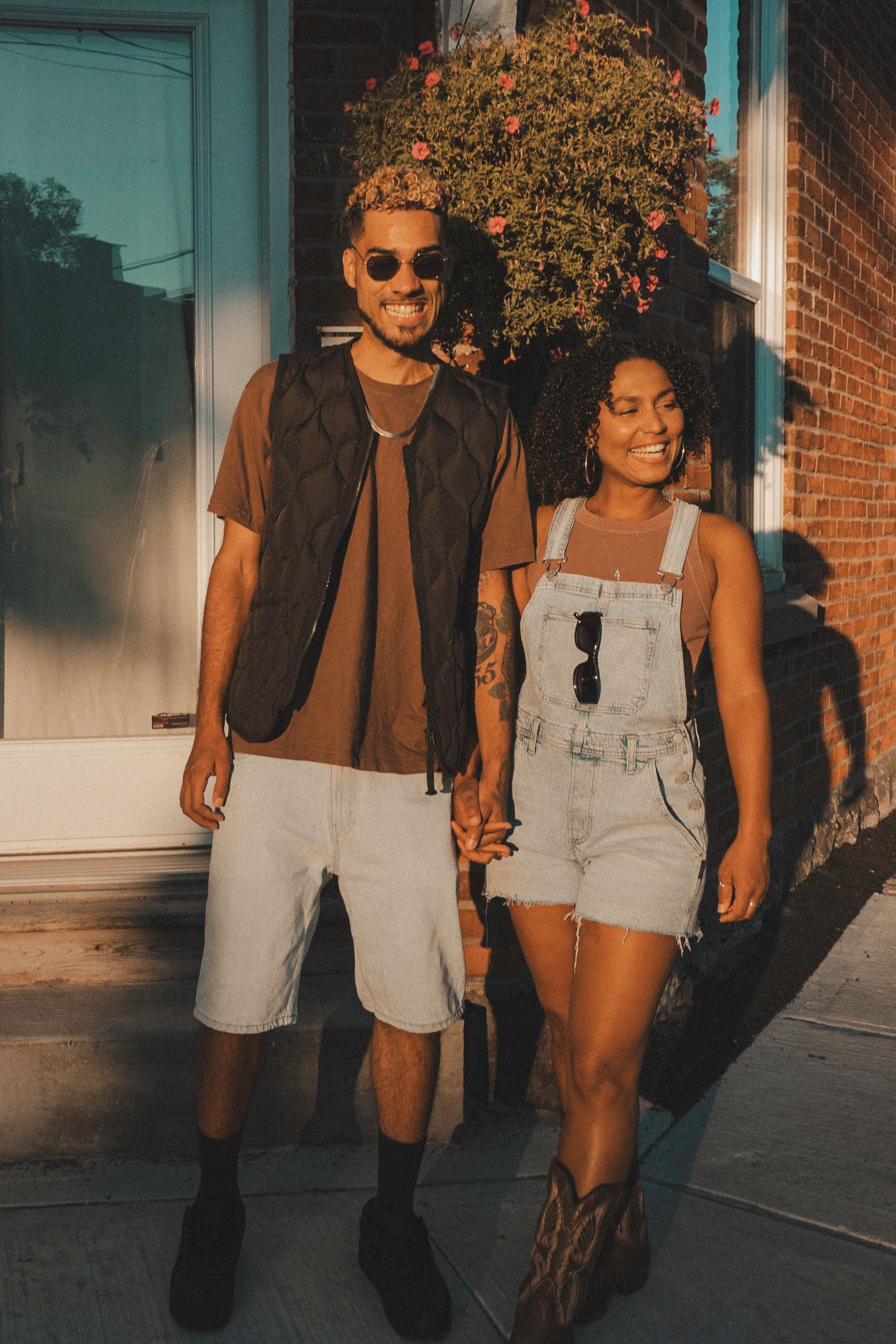 Couple smiling, holding hands. Man in vest, shorts; woman in overalls, boots, standing by building.