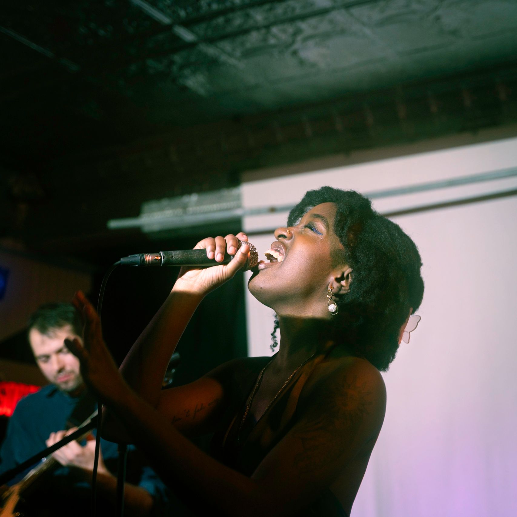 A woman singing into a microphone onstage; a man plays guitar in the background.